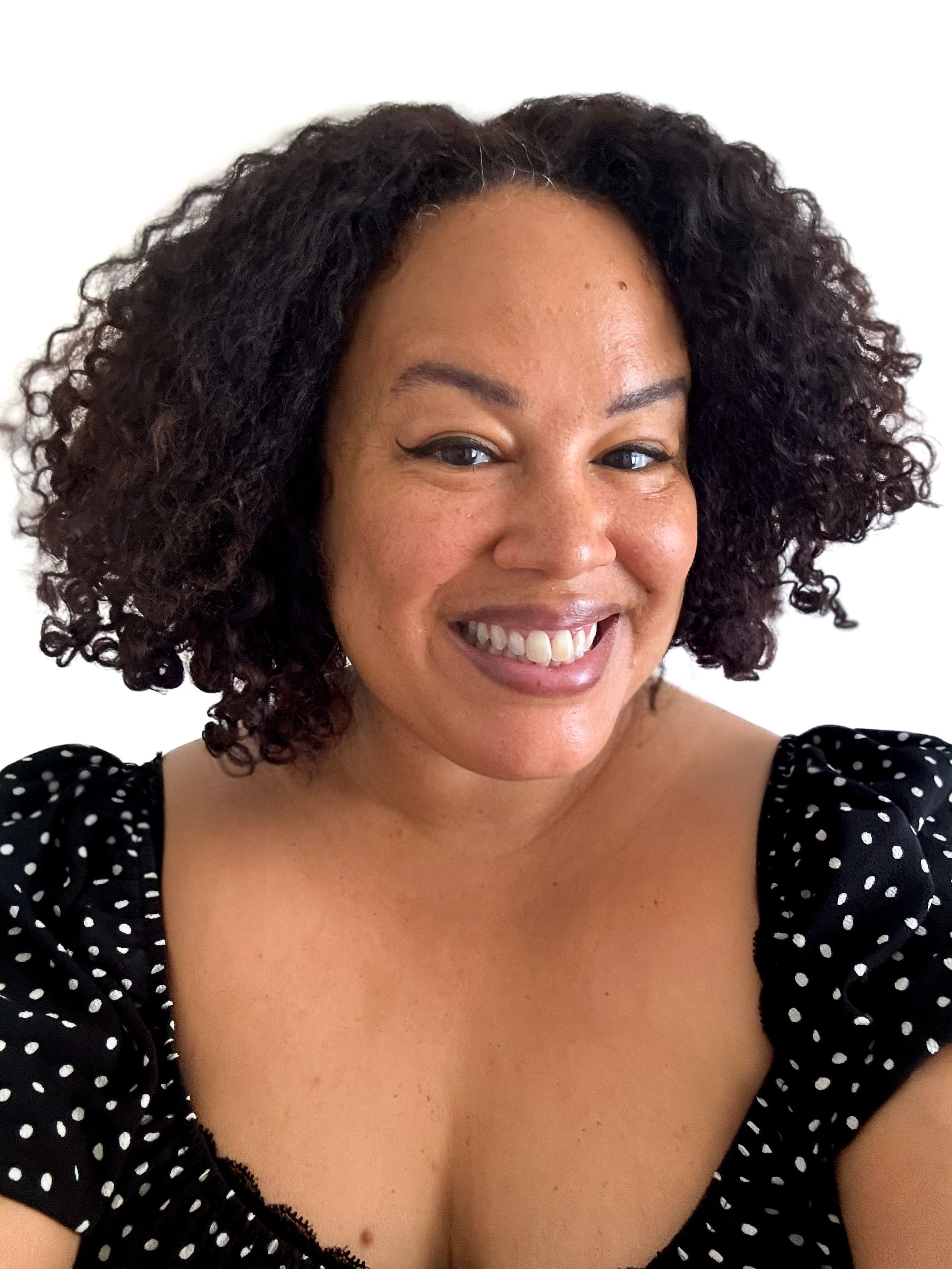 A smiling woman with dark curly hair wears a black and white polka dot top.