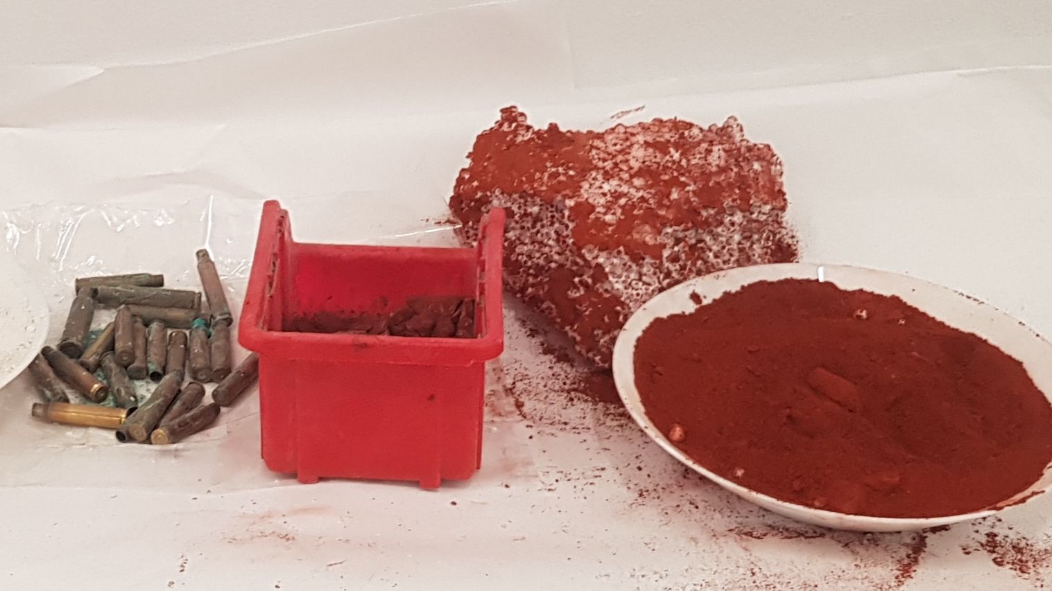Spent bullet casings, reddish-brown powder in a red container and white dish, and a large reddish lump with white crystals, likely an explosive.