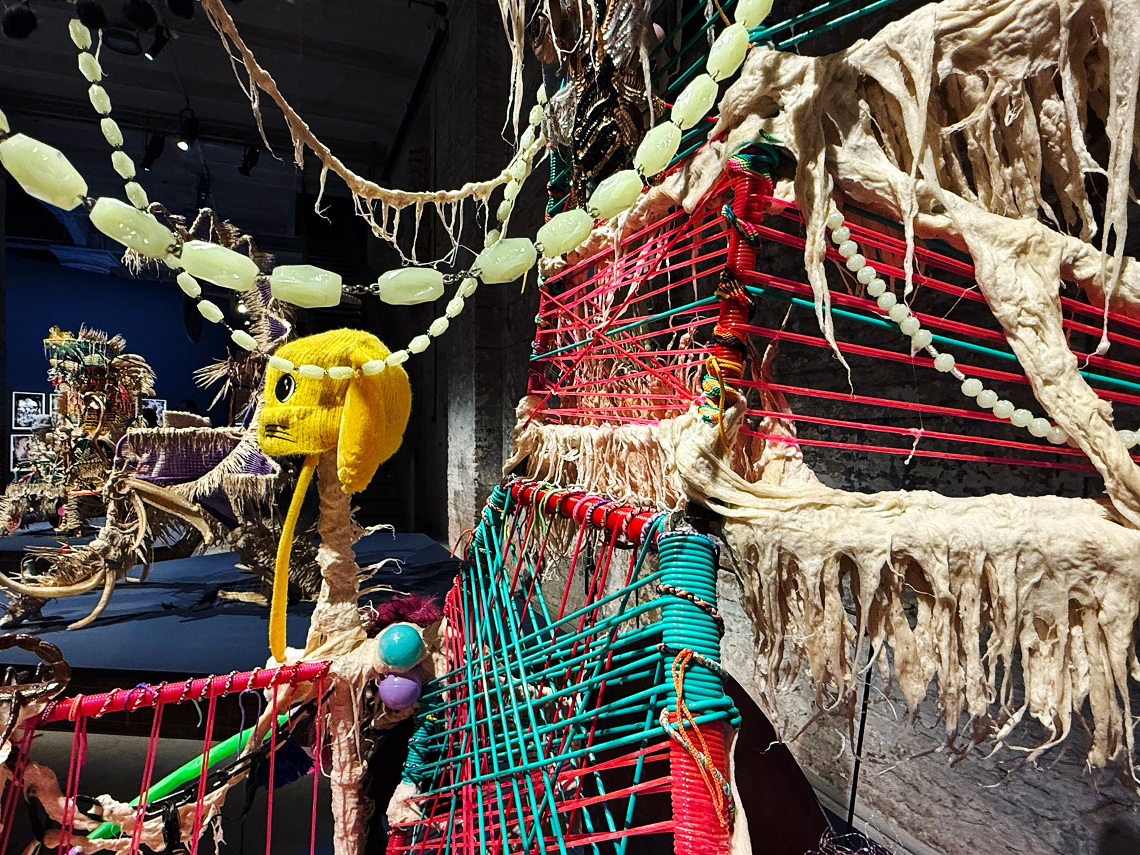 A vibrant mixed-media art installation featuring pink and teal woven sections, textured beige fibers, a yellow cartoon-like head, and beaded strands.