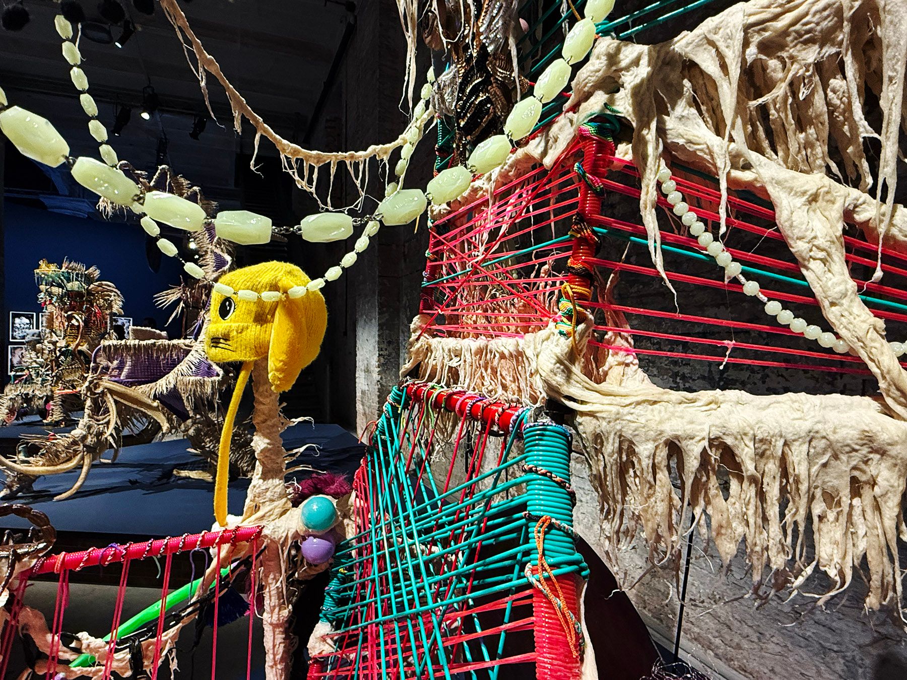 A vibrant mixed-media art installation featuring pink and teal woven sections, textured beige fibers, a yellow cartoon-like head, and beaded strands.