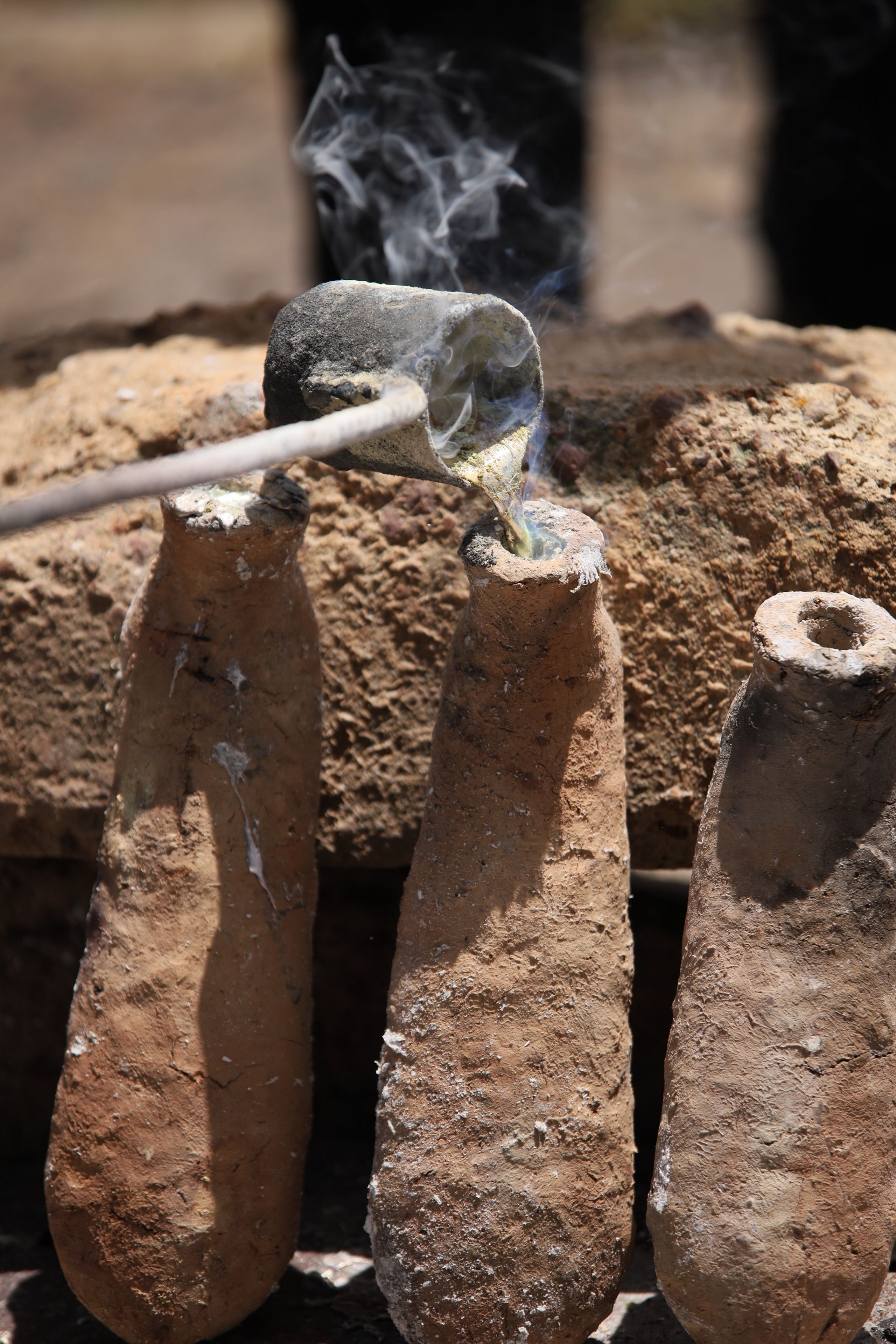 Molten metal pours from a crucible into a tall, conical clay mold, emitting smoke.