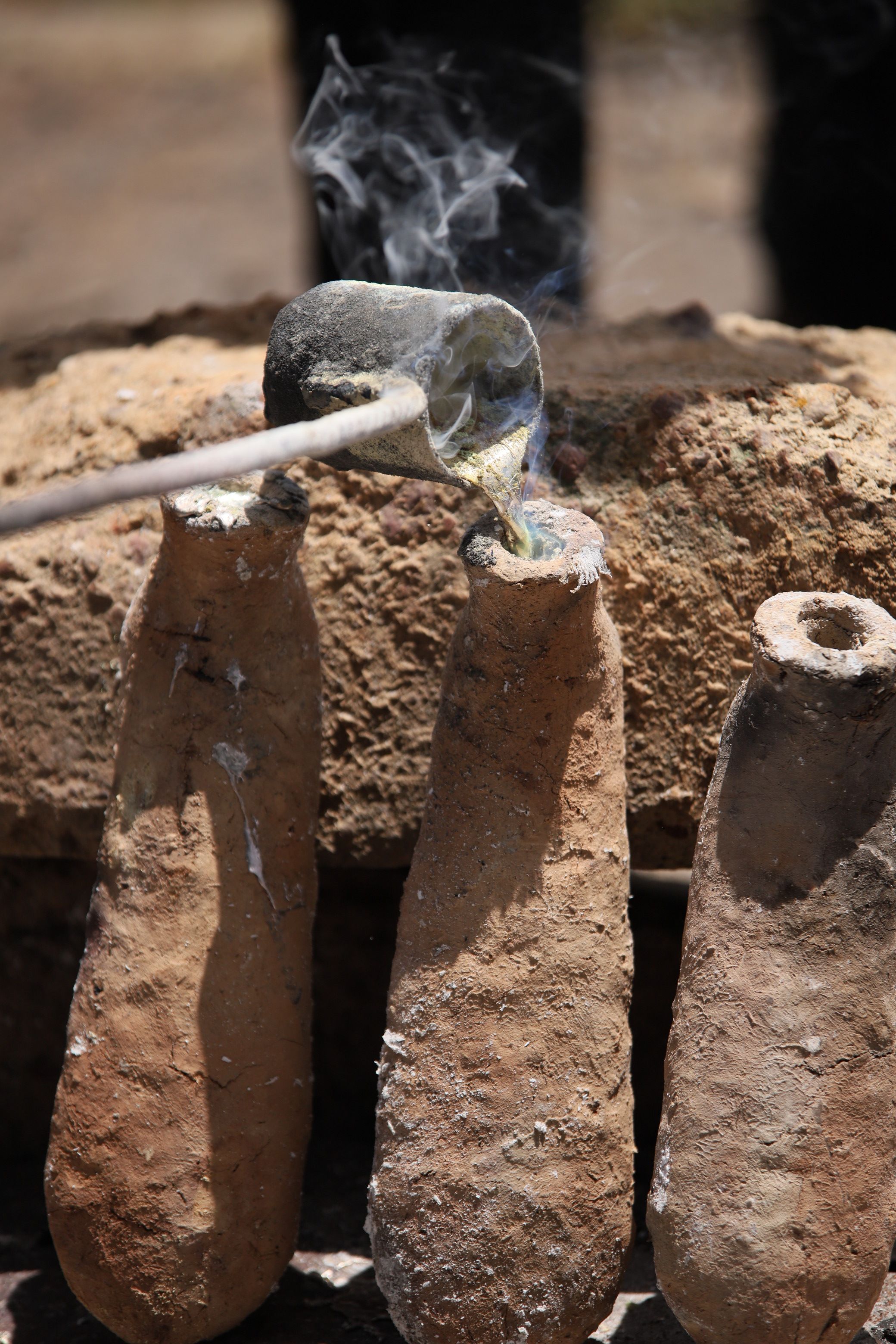 Molten metal pours from a crucible into a tall, conical clay mold, emitting smoke.