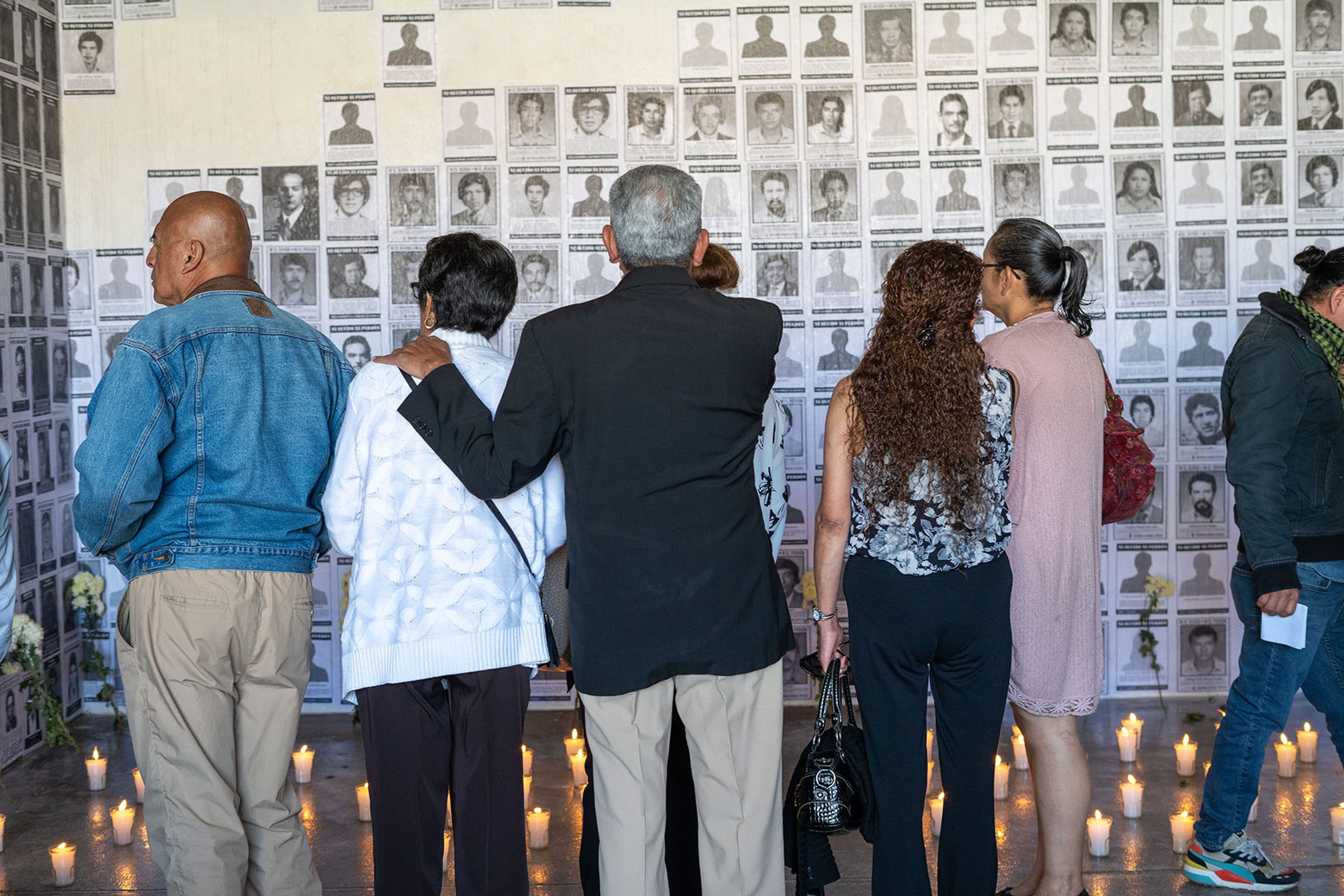 A group of people stand facing a wall covered with black and white portraits, with lit candles on the floor.