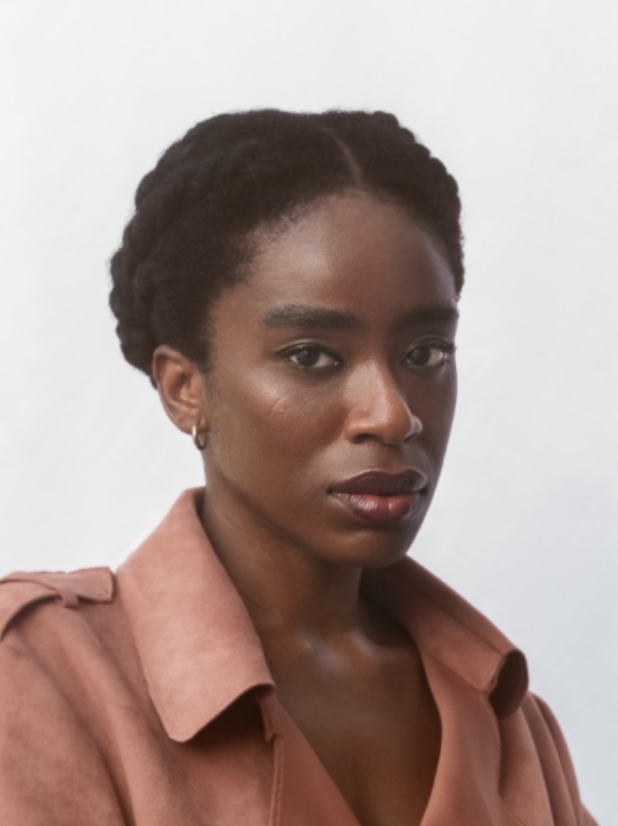 A Black woman with dark braided hair and a neutral expression wears a rose-brown jacket.