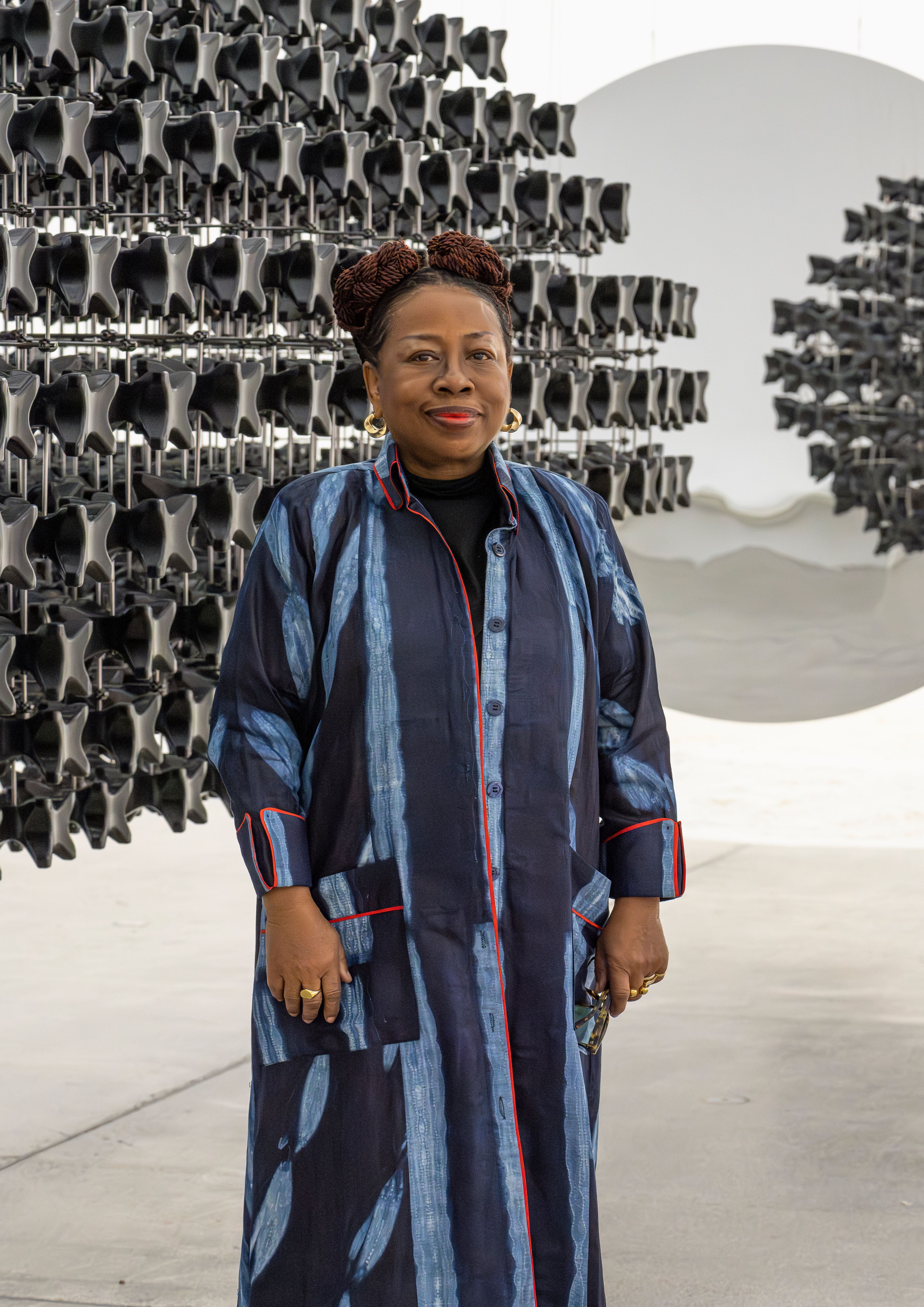 A smiling Black woman in a long blue patterned dress stands before a dark geometric art installation and a large white disc.