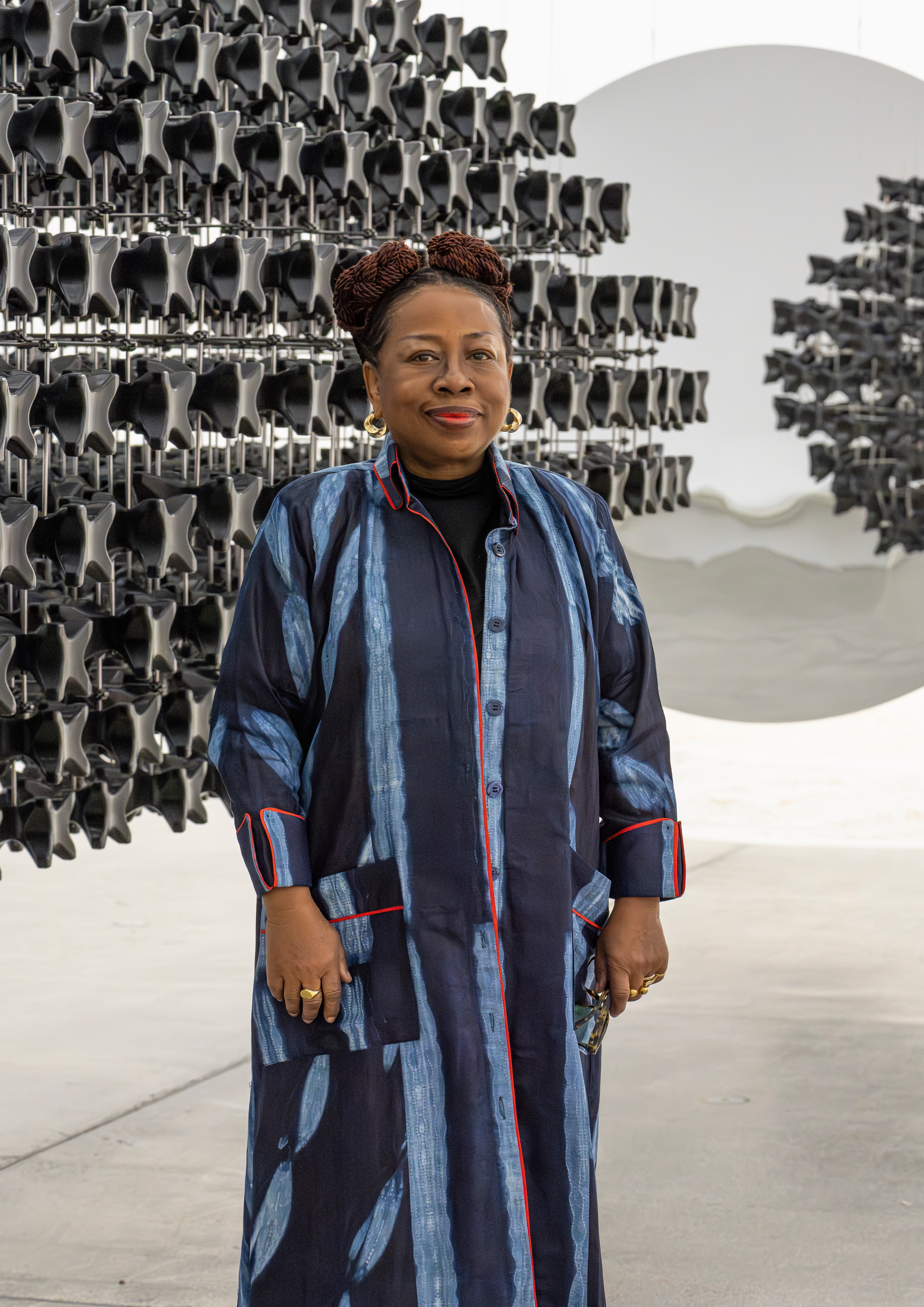 A smiling Black woman in a long blue patterned dress stands before a dark geometric art installation and a large white disc.