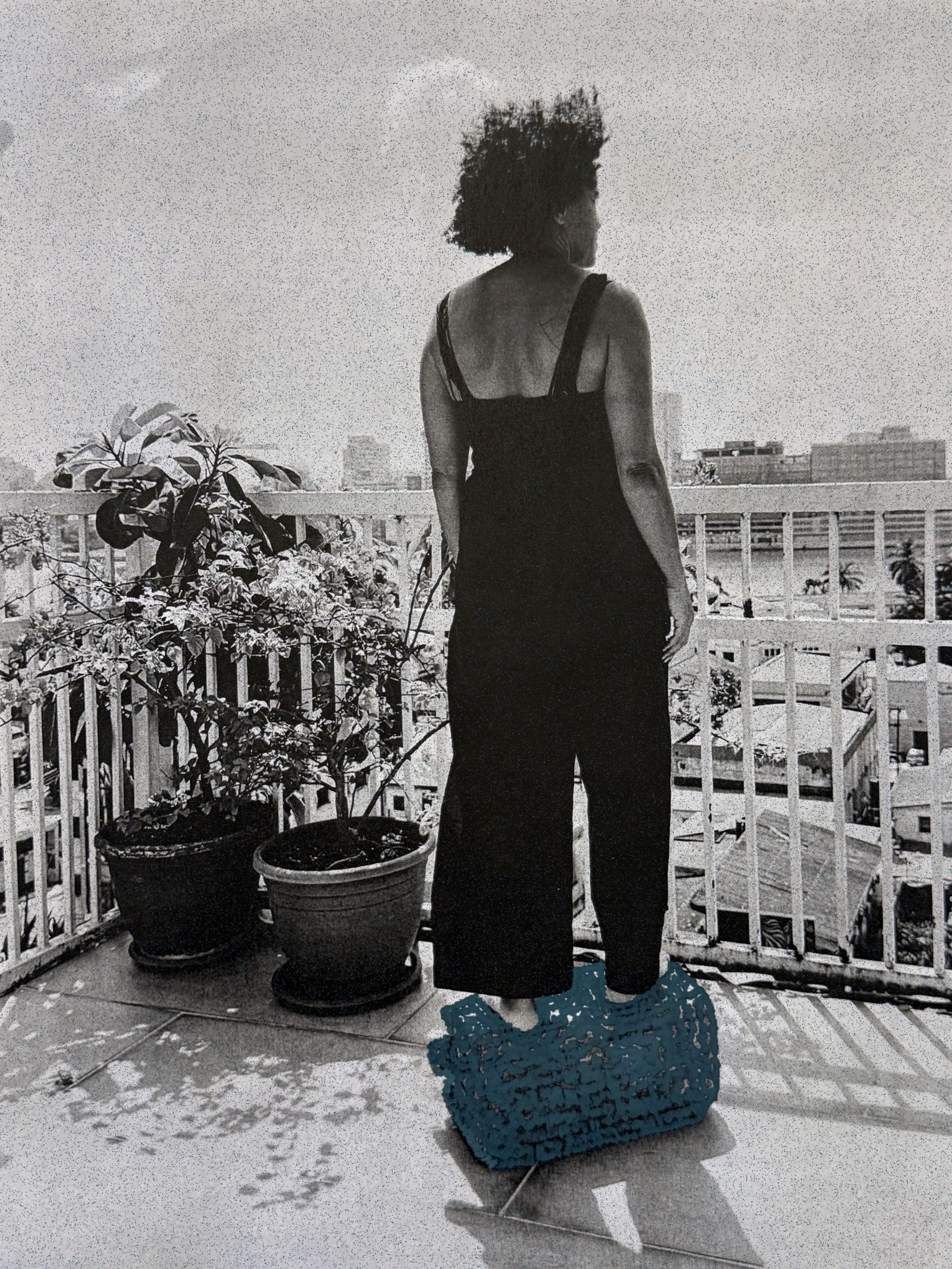 Grainy monochrome image of a person in a black jumpsuit standing on a teal woven object on a balcony, looking at a city skyline.