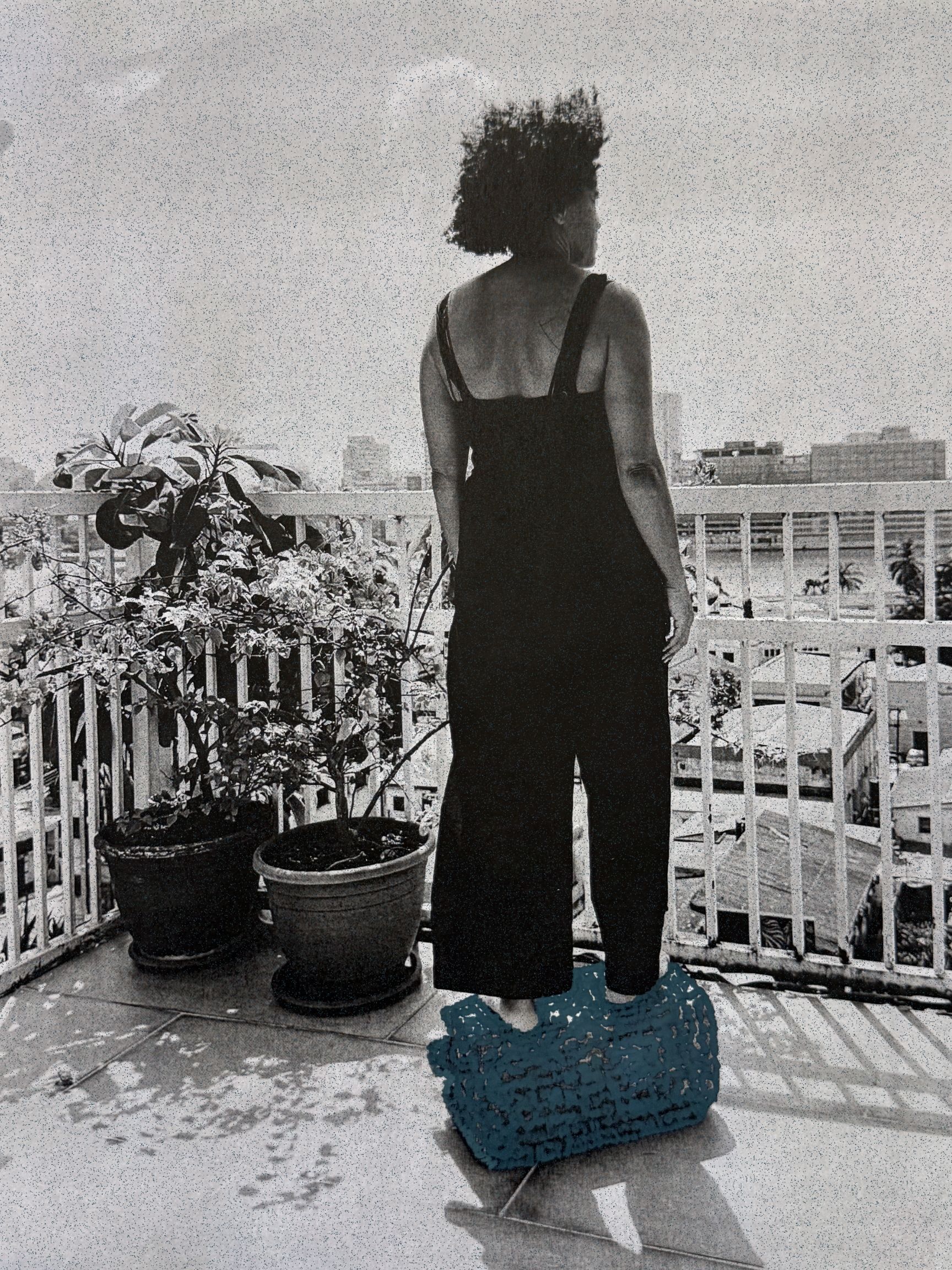 Grainy monochrome image of a person in a black jumpsuit standing on a teal woven object on a balcony, looking at a city skyline.