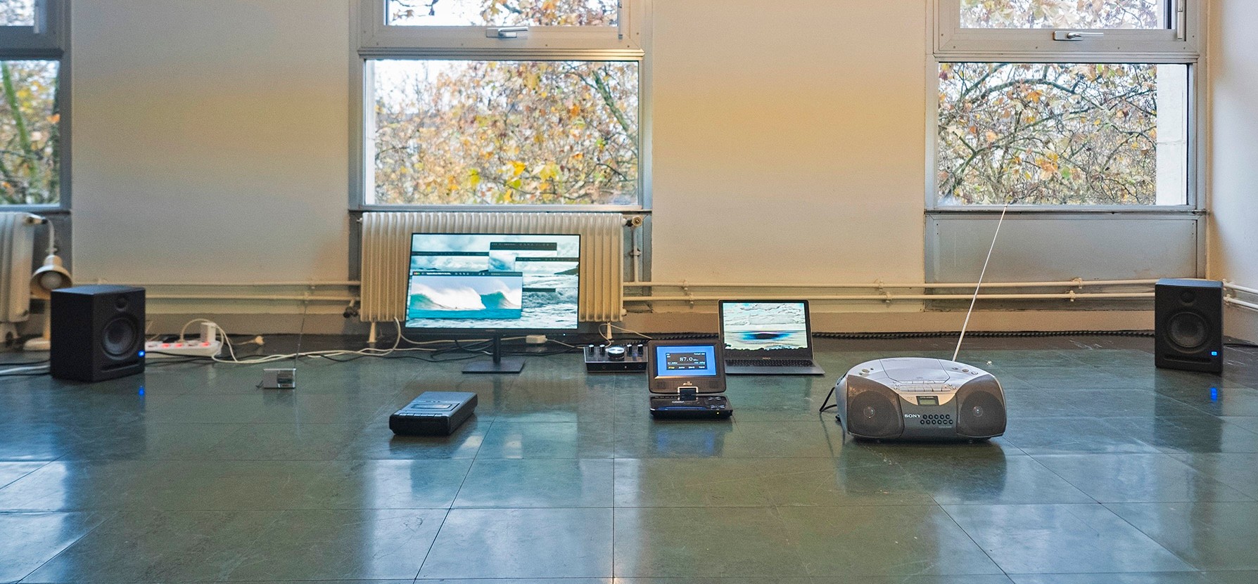 Various electronic devices, including a glitched monitor, two laptops, a boombox, and two speakers, are spread on a tiled floor in a room with large windows overlooking trees.