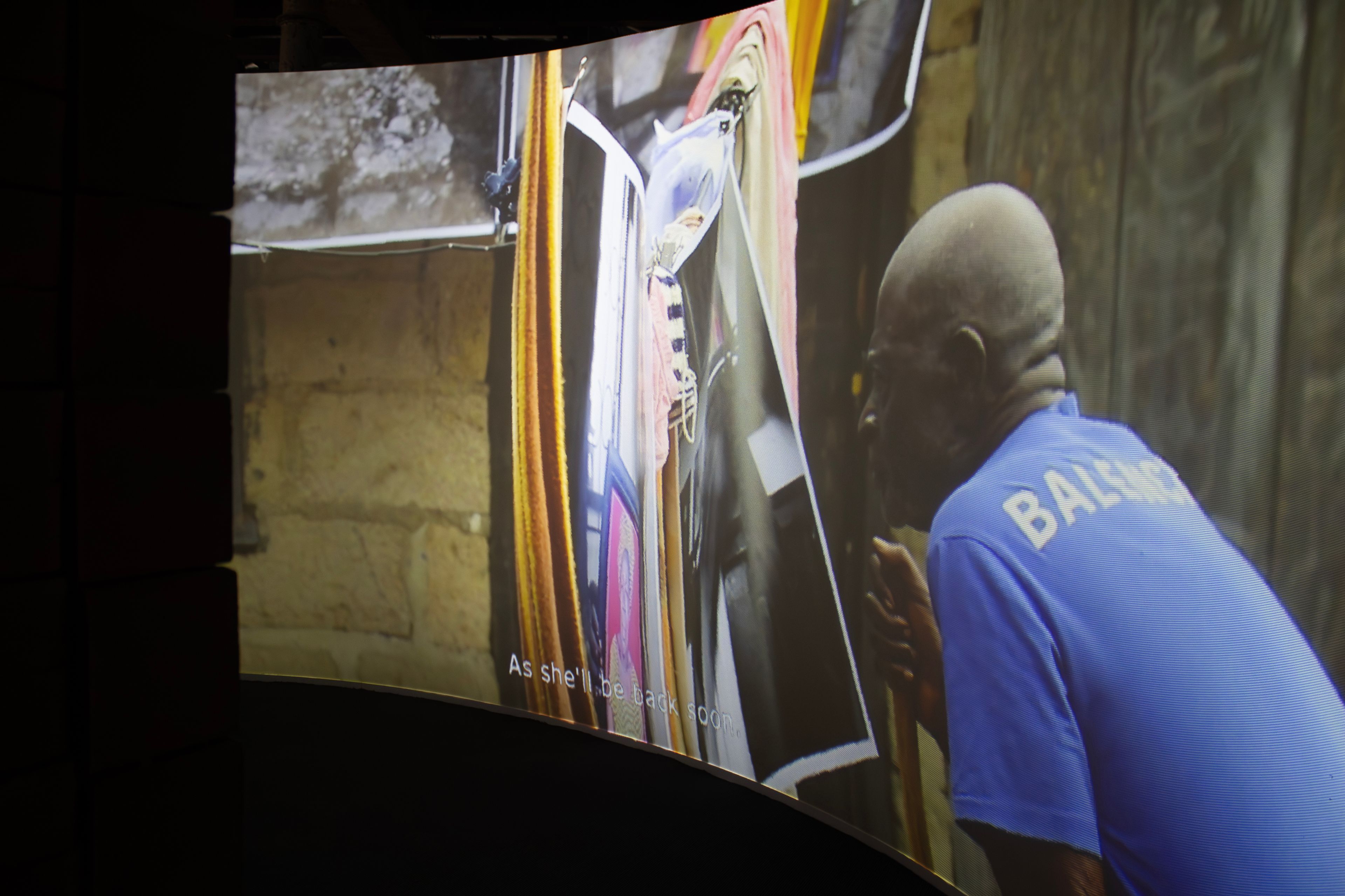 A curved screen displays an older Black man, seen from behind, wearing a blue Balenciaga shirt and looking at colorful hanging fabrics.
