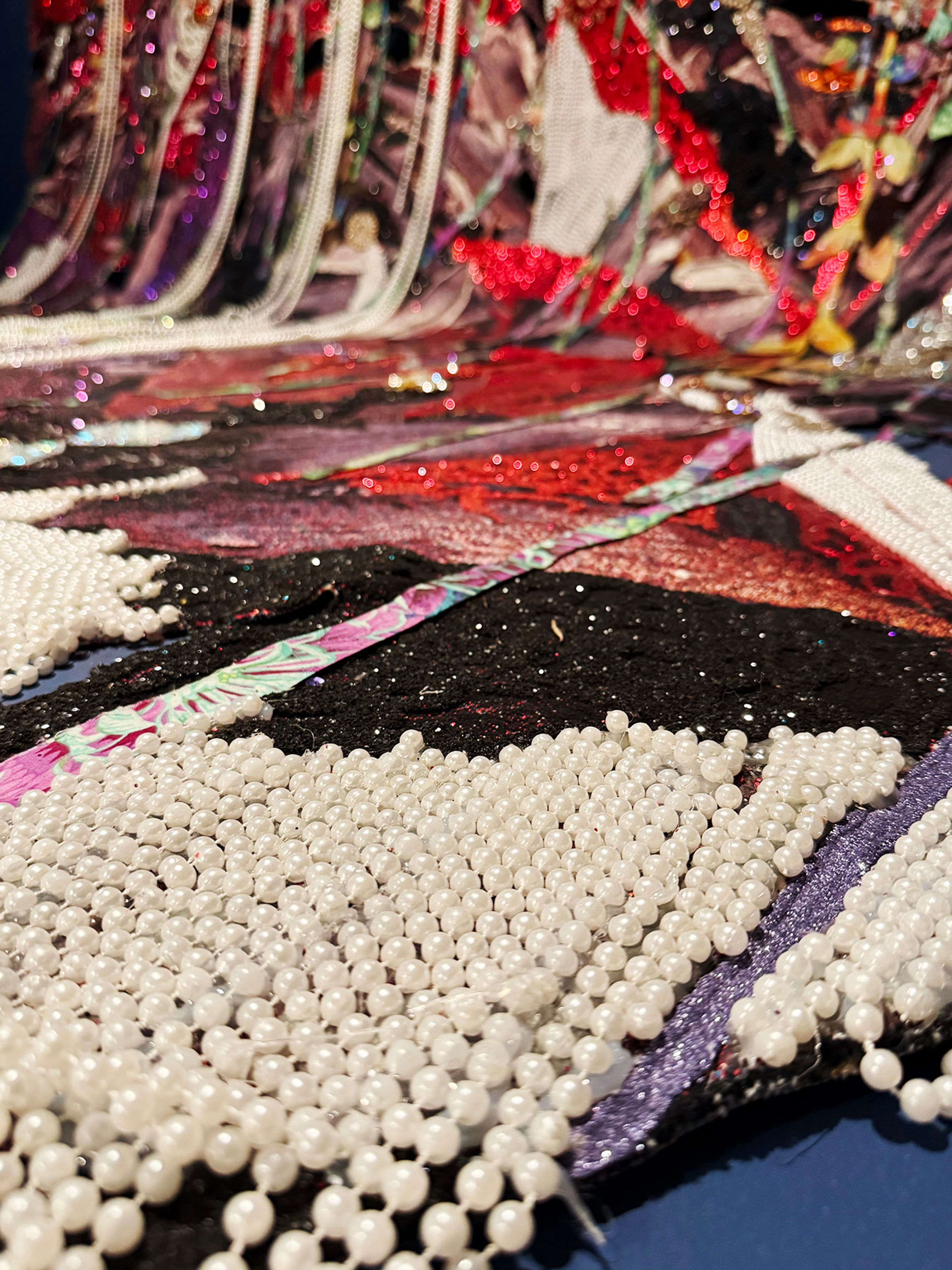 Close-up of an elaborate textile art piece, featuring white pearl fringe in the foreground and a blurred background of sparkling red, black, and patterned fabrics.