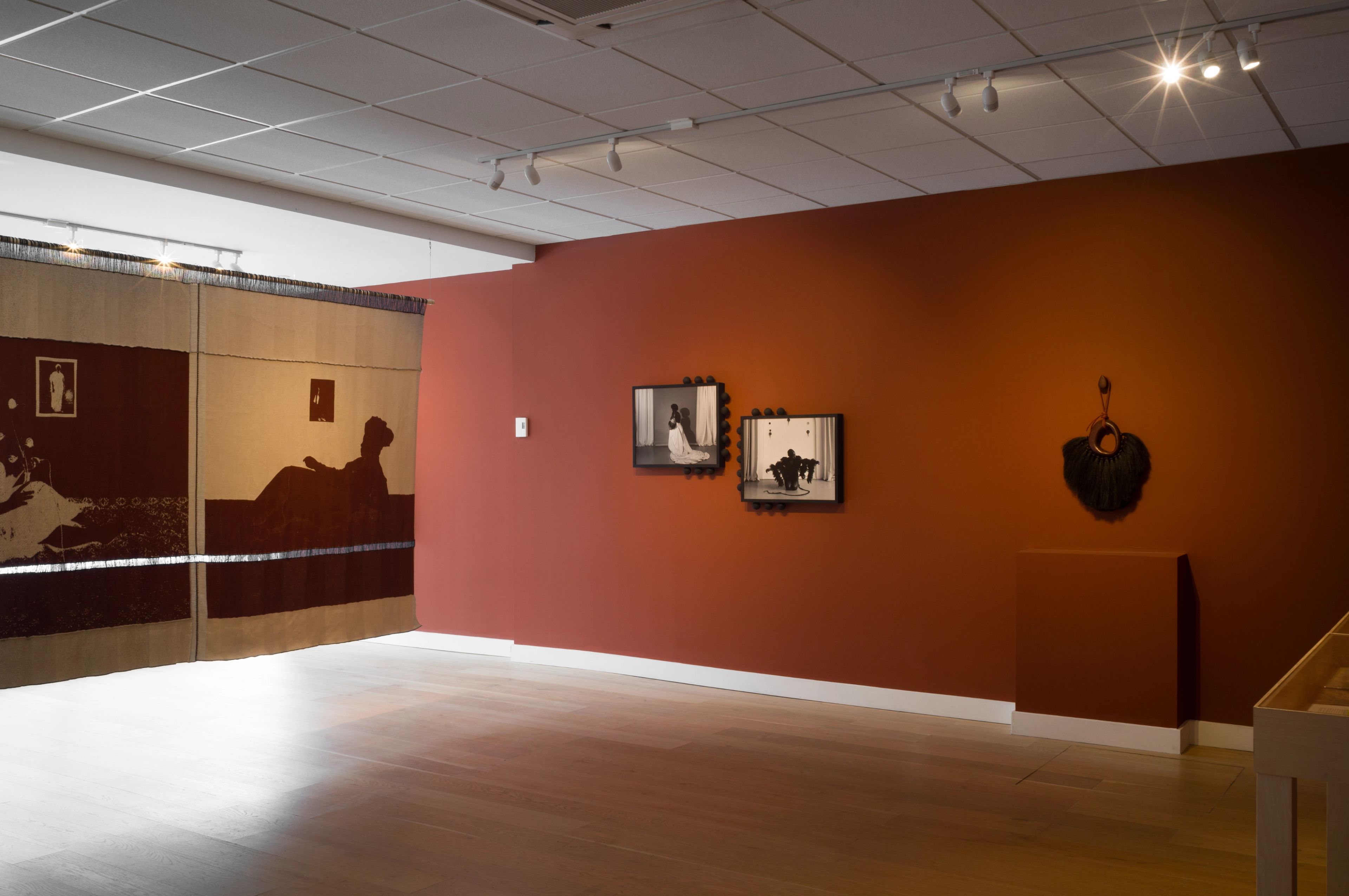 A gallery space featuring a large tapestry, a reddish-brown wall with two framed photographs, and a dark, fan-like sculpture.