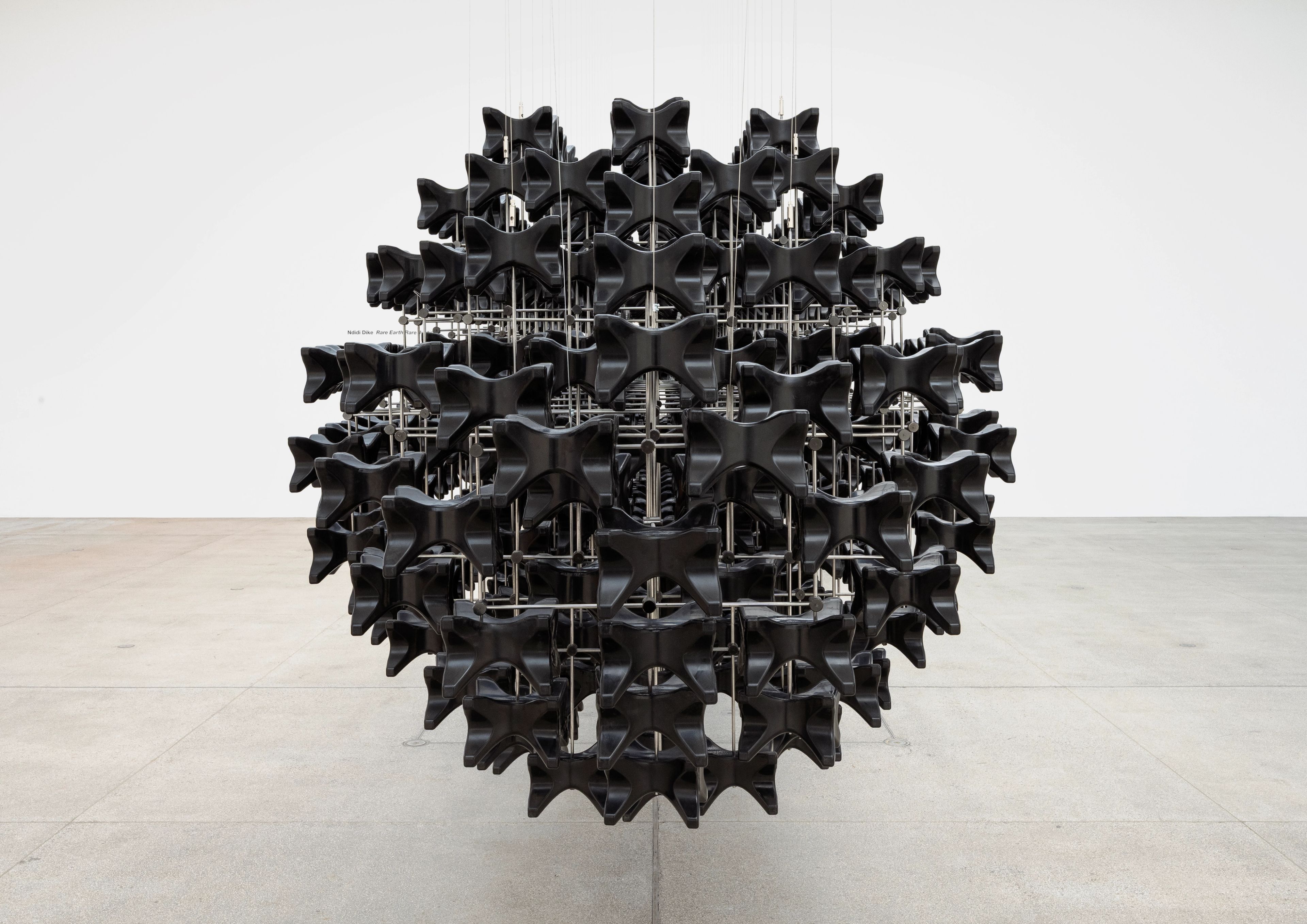 A large, black, spherical sculpture made of repeating bow-tie-shaped modules on a metal grid, suspended in a white gallery.