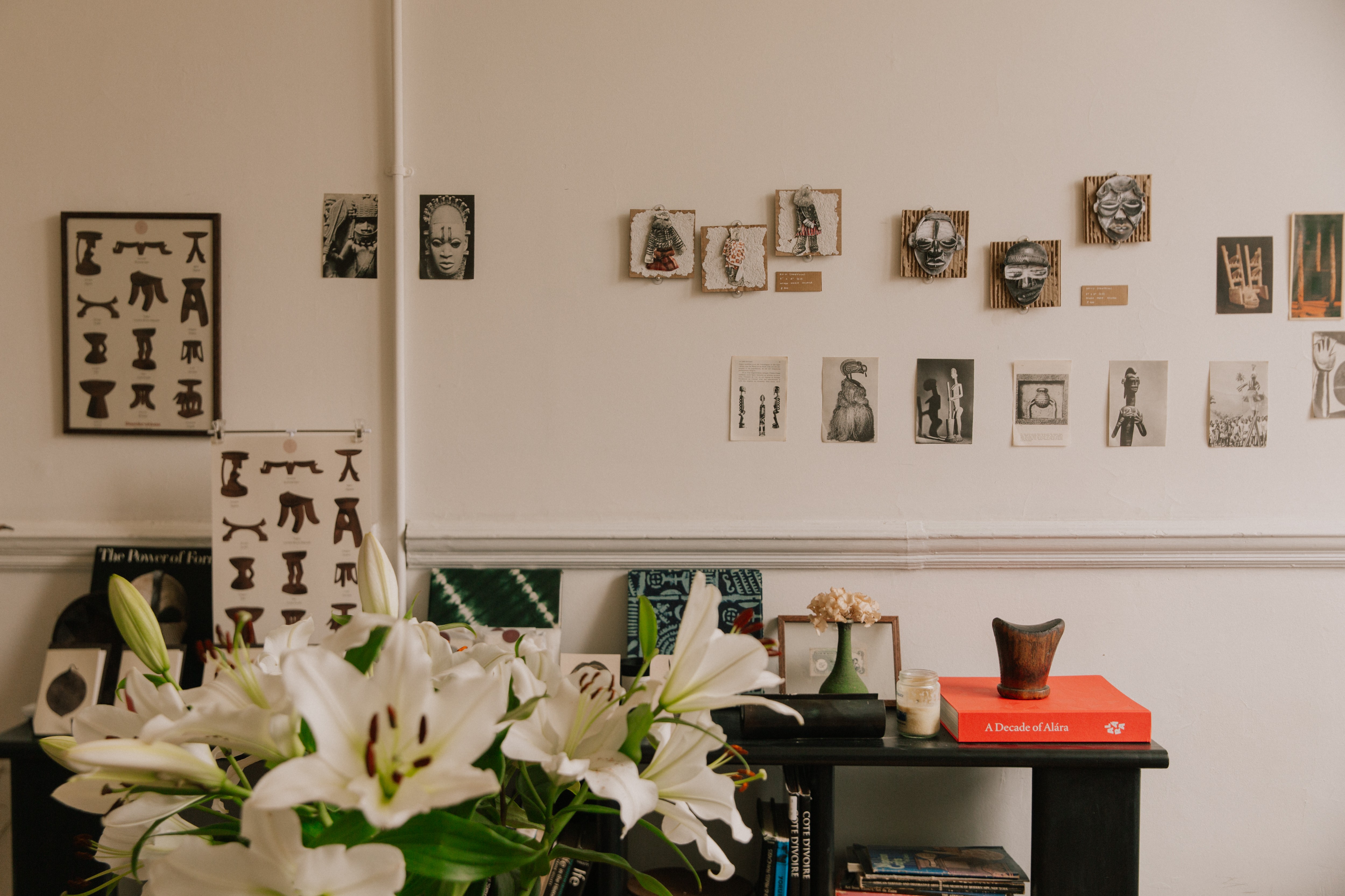 A bouquet of white lilies in the foreground, with a console table and a white wall decorated with an array of diverse artworks and prints, some featuring masks and figures.