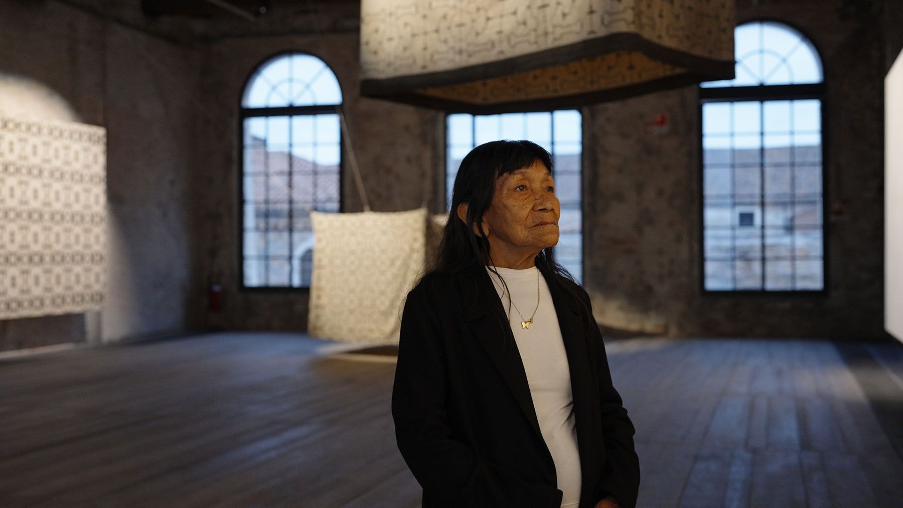 An older Indigenous woman stands in an art gallery with patterned textiles and large arched windows.