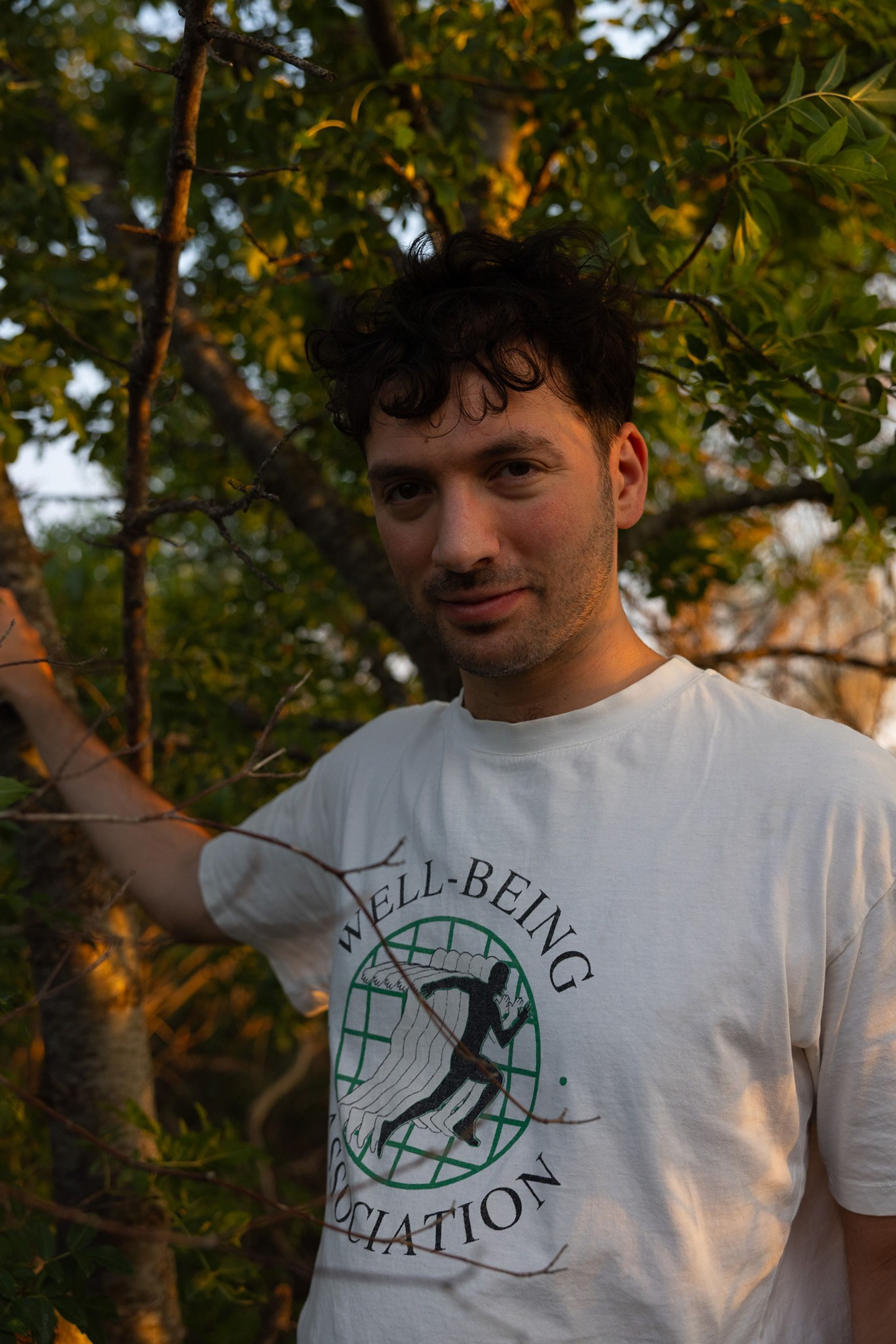 A man with dark curly hair and a slight beard, wearing a white "WELL-BEING ASSOCIATION" t-shirt, stands among trees in warm sunlight.