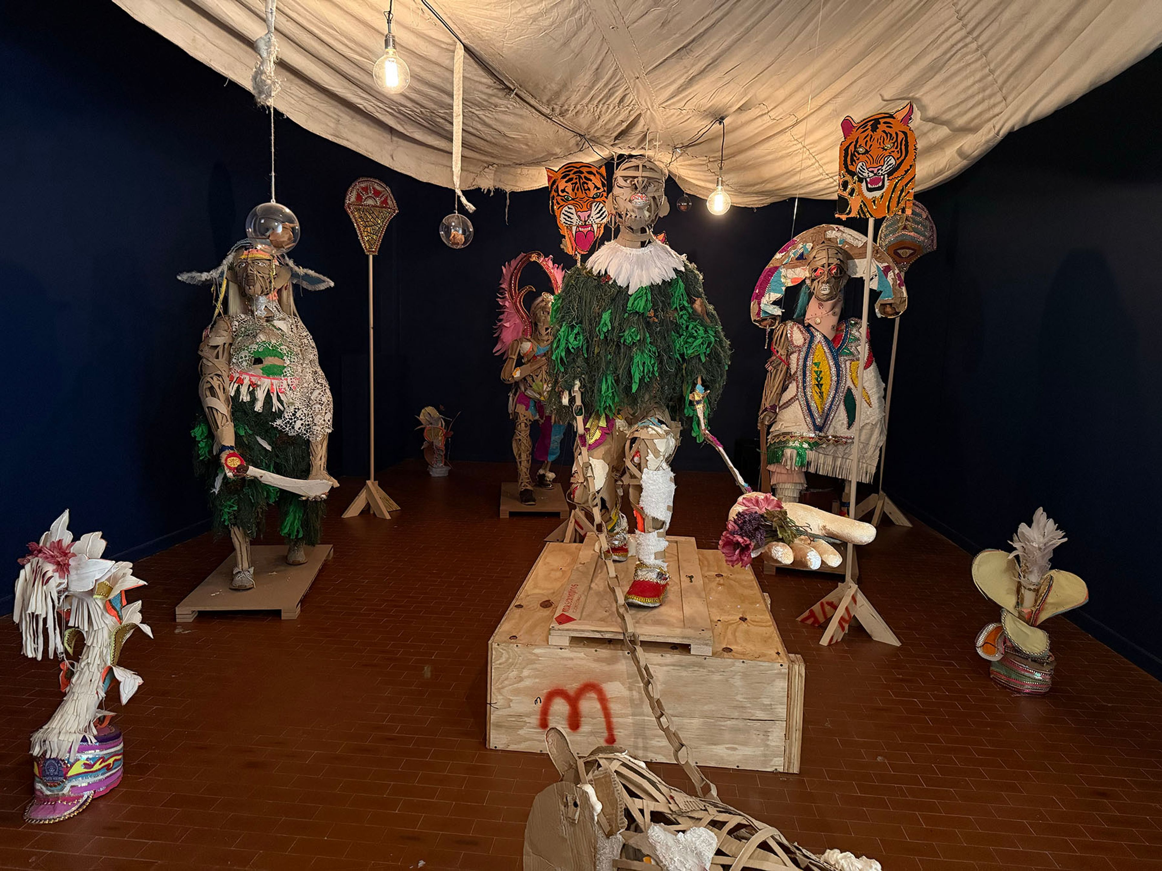 An art exhibition featuring multiple intricately decorated, mixed-media figures with masks, some resembling tigers, under a draped white ceiling.