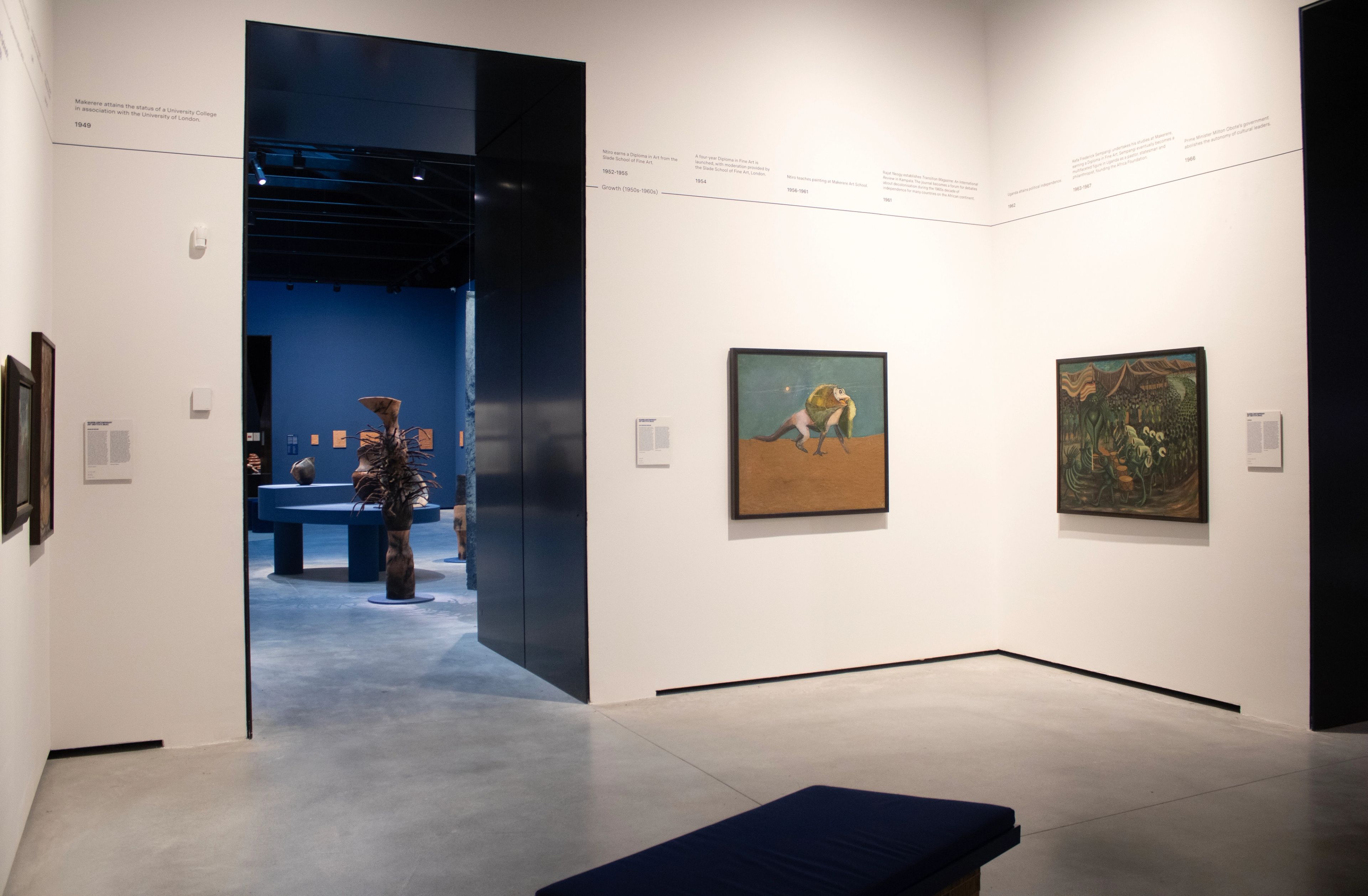 A white-walled art gallery with a concrete floor, an open doorway leading to a dark blue room with sculptures, two paintings on the right wall, a timeline along the top, and a blue bench in the foreground.