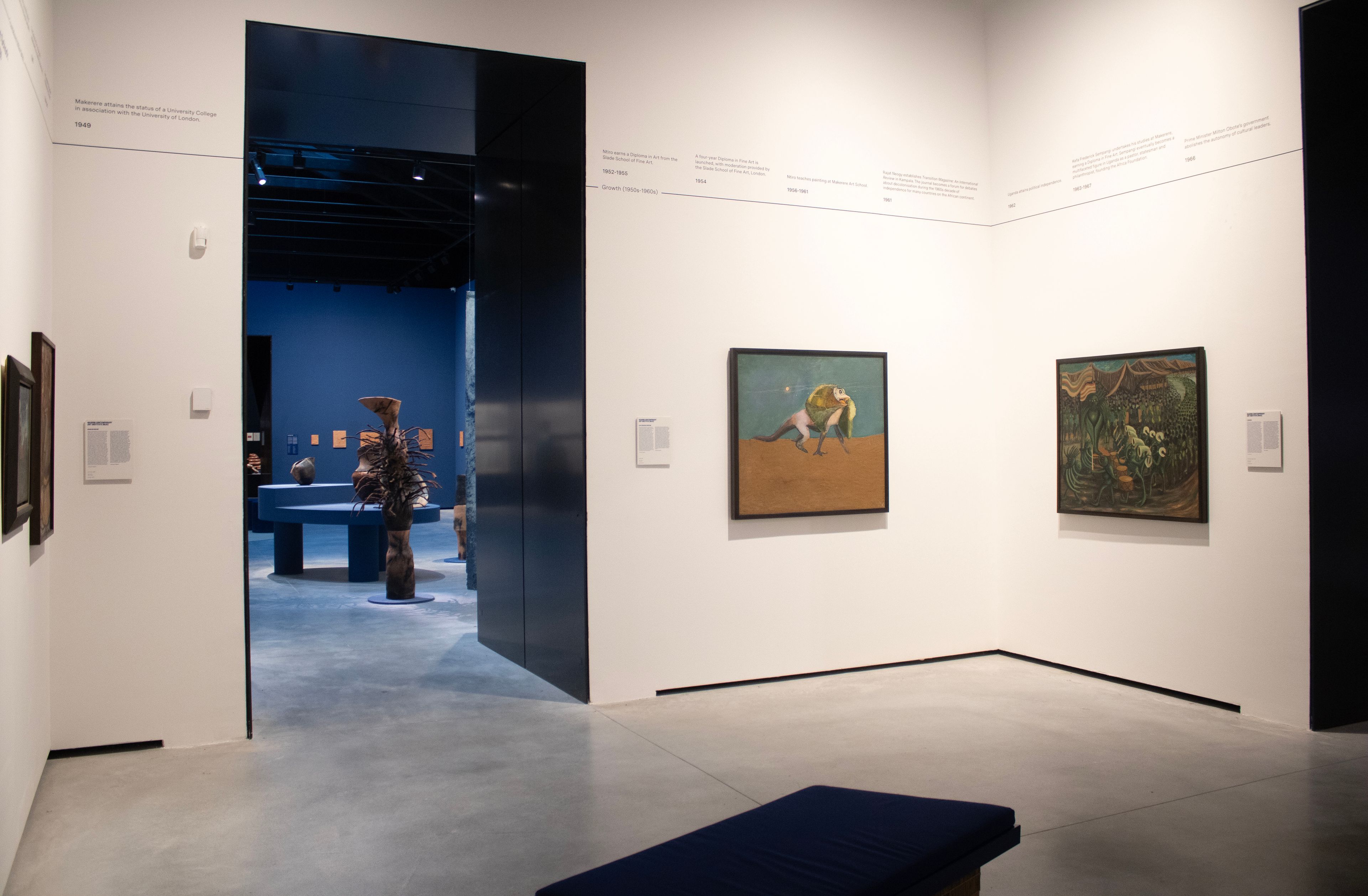 A white-walled art gallery with a concrete floor, an open doorway leading to a dark blue room with sculptures, two paintings on the right wall, a timeline along the top, and a blue bench in the foreground.