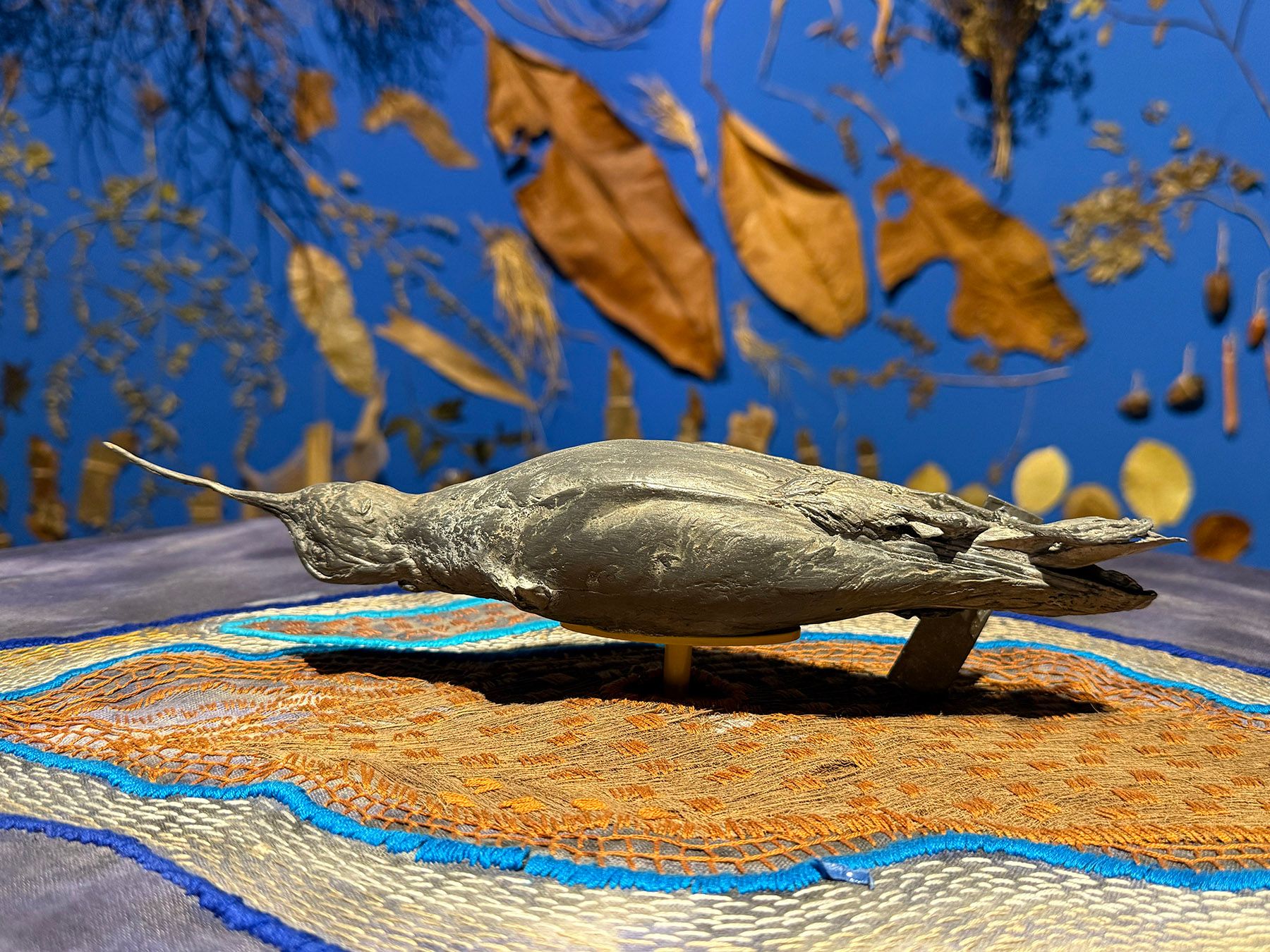 A grey taxidermied bird with a long beak lies on an embroidered cloth, against a blue wall decorated with dried leaves.