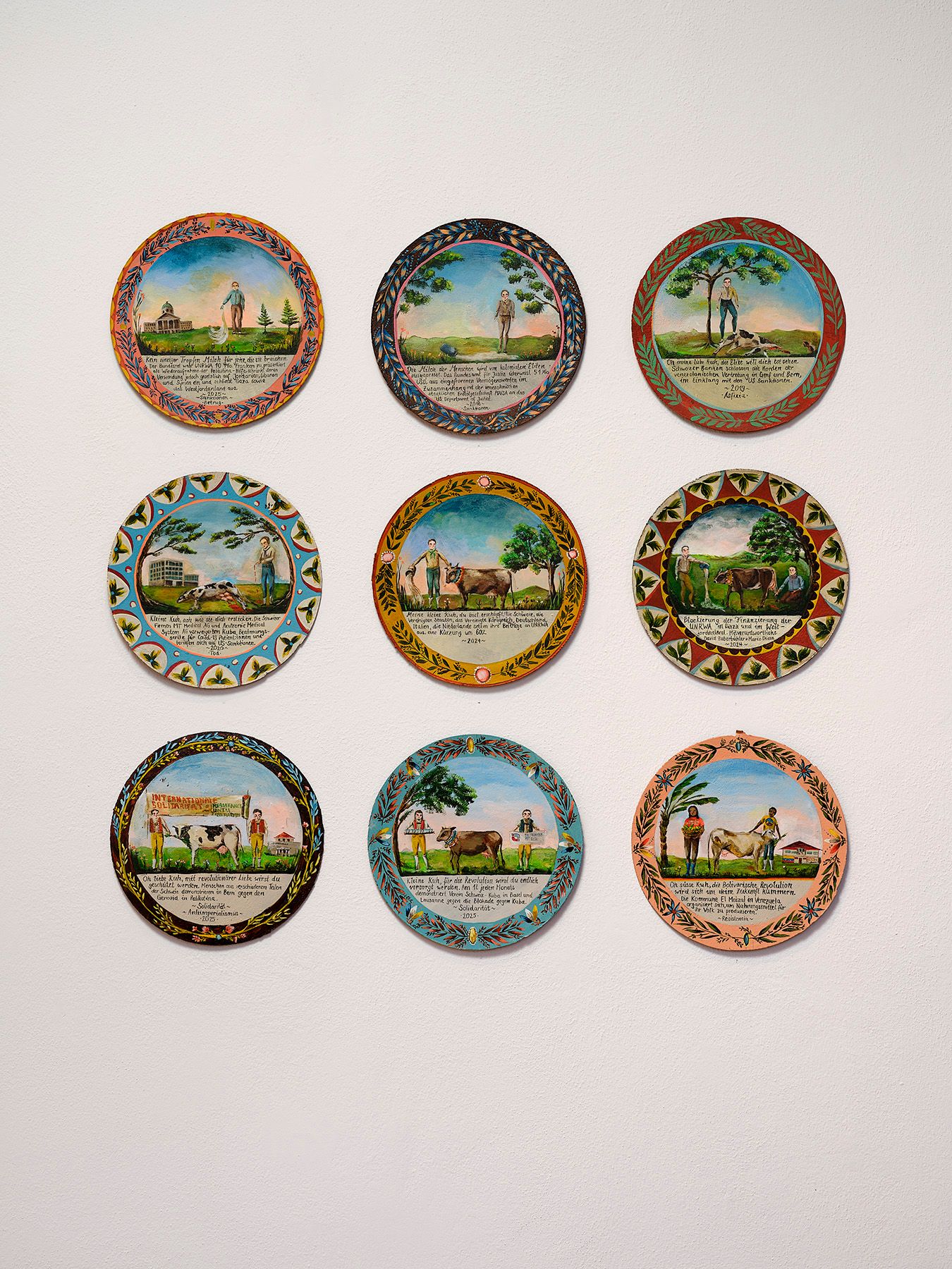 Nine circular folk art paintings, depicting people, animals, and text, arranged in a 3x3 grid on a white wall.