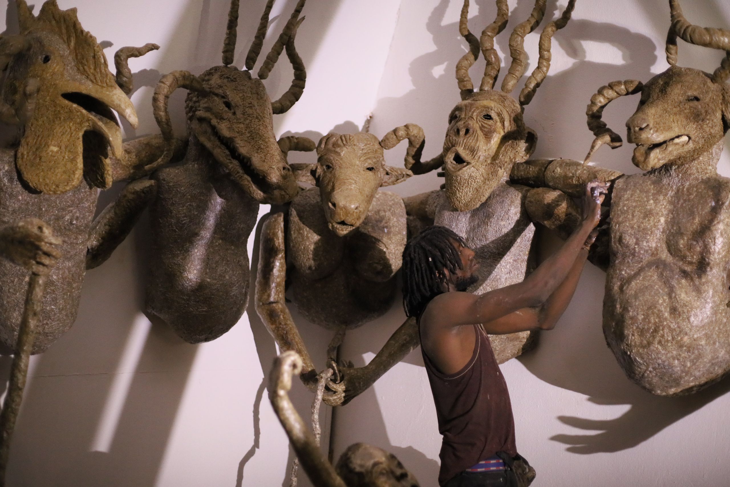 An artist adjusts one of five large, textured sculptures of human figures with animal heads mounted on a white wall.