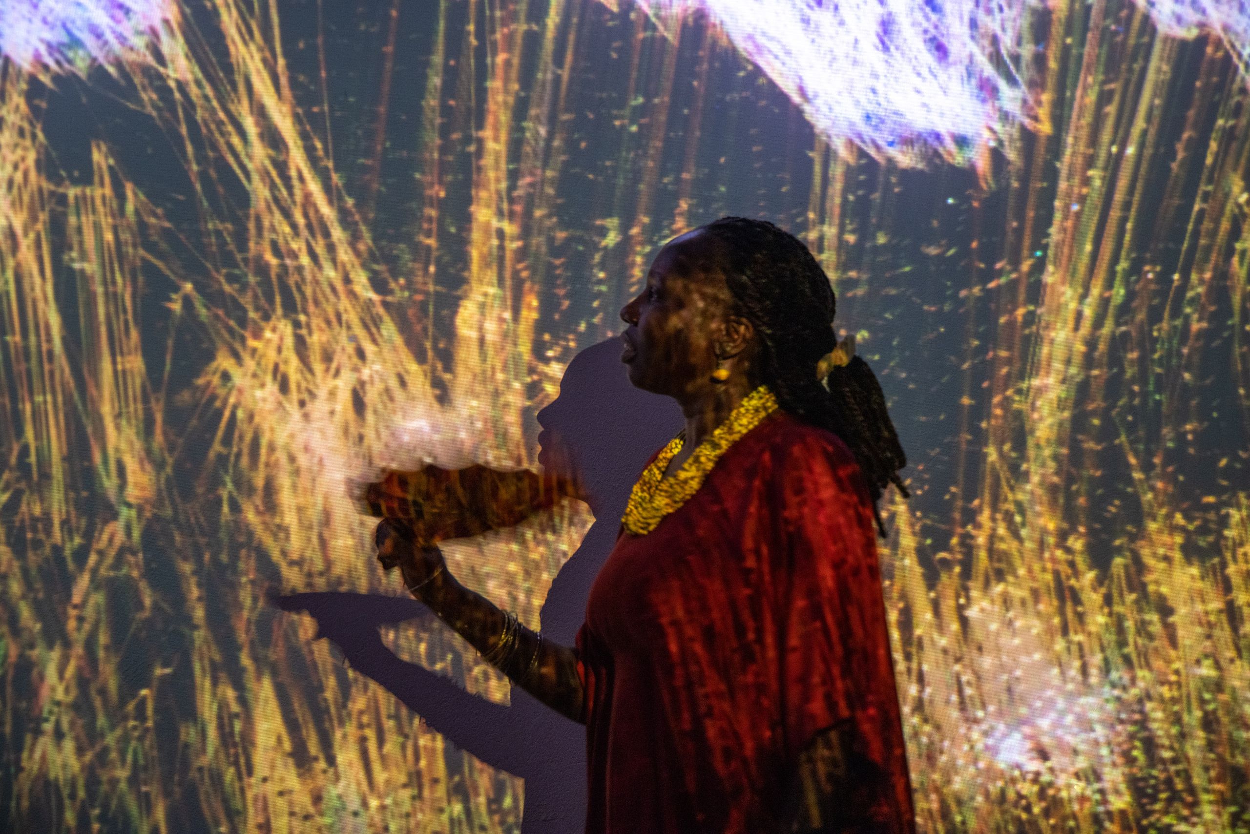 A woman in a red top and gold necklace stands in profile, illuminated by shimmering golden and purple light projections on a wall.