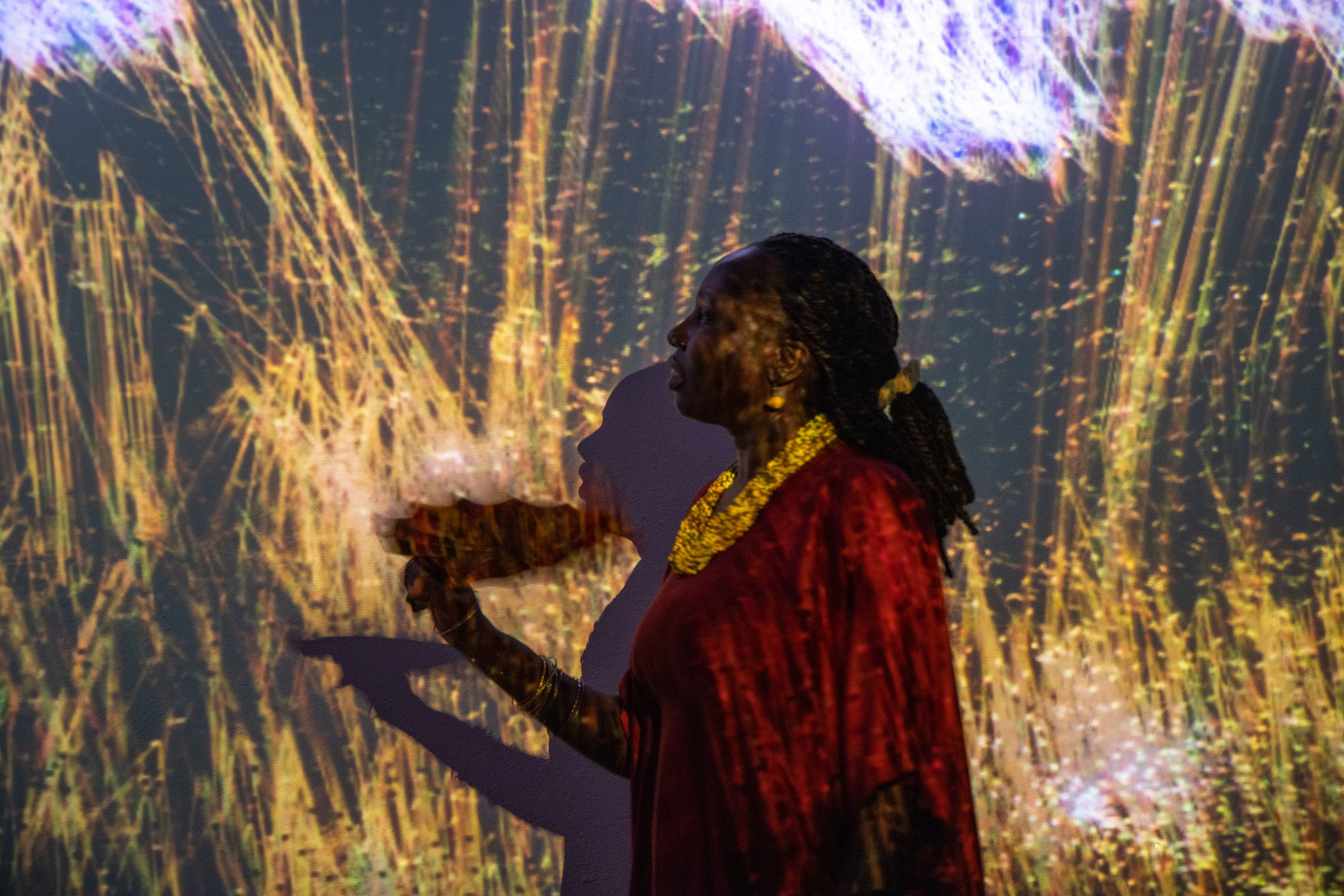 A woman in a red top and gold necklace stands in profile, illuminated by shimmering golden and purple light projections on a wall.