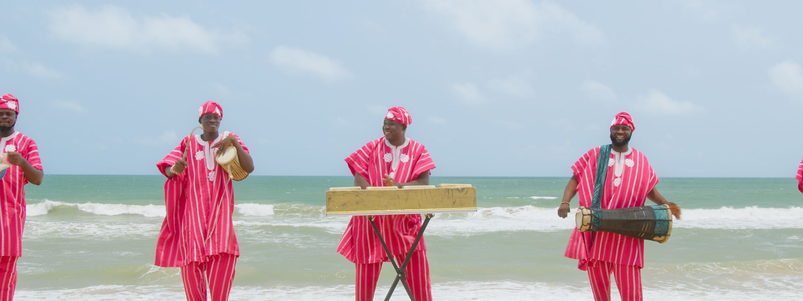 A group of musicians in traditional pink and white striped outfits playing instruments on a beach.