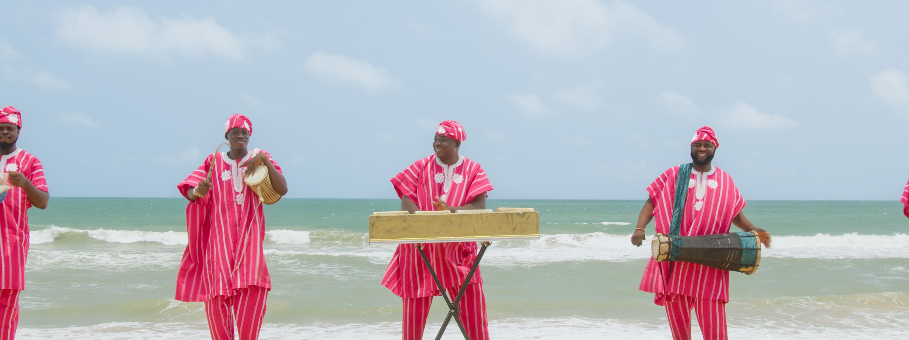 A group of musicians in traditional pink and white striped outfits playing instruments on a beach.