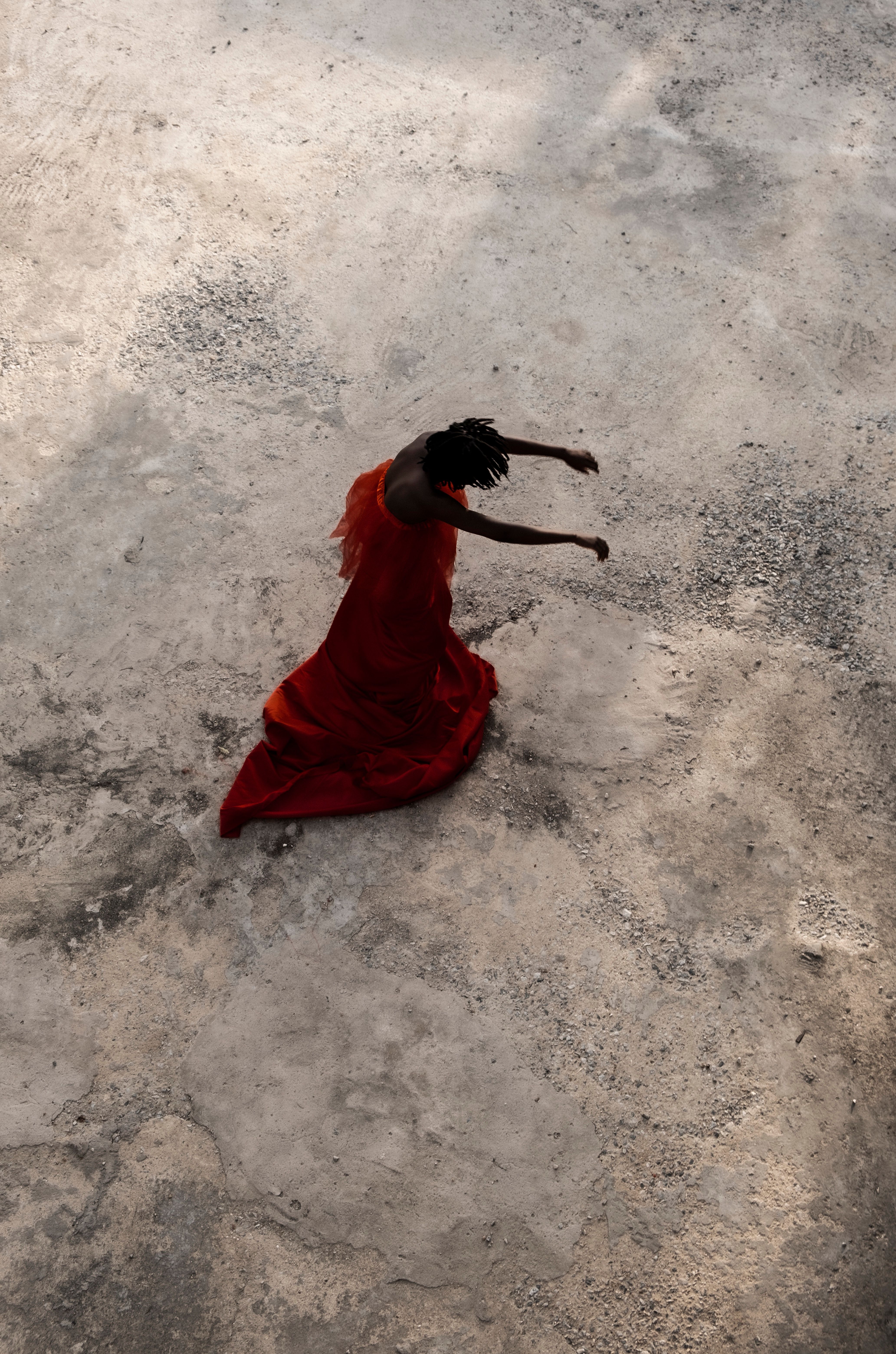 An overhead view of a person in a flowing orange dress dancing on a textured, light-colored ground.
