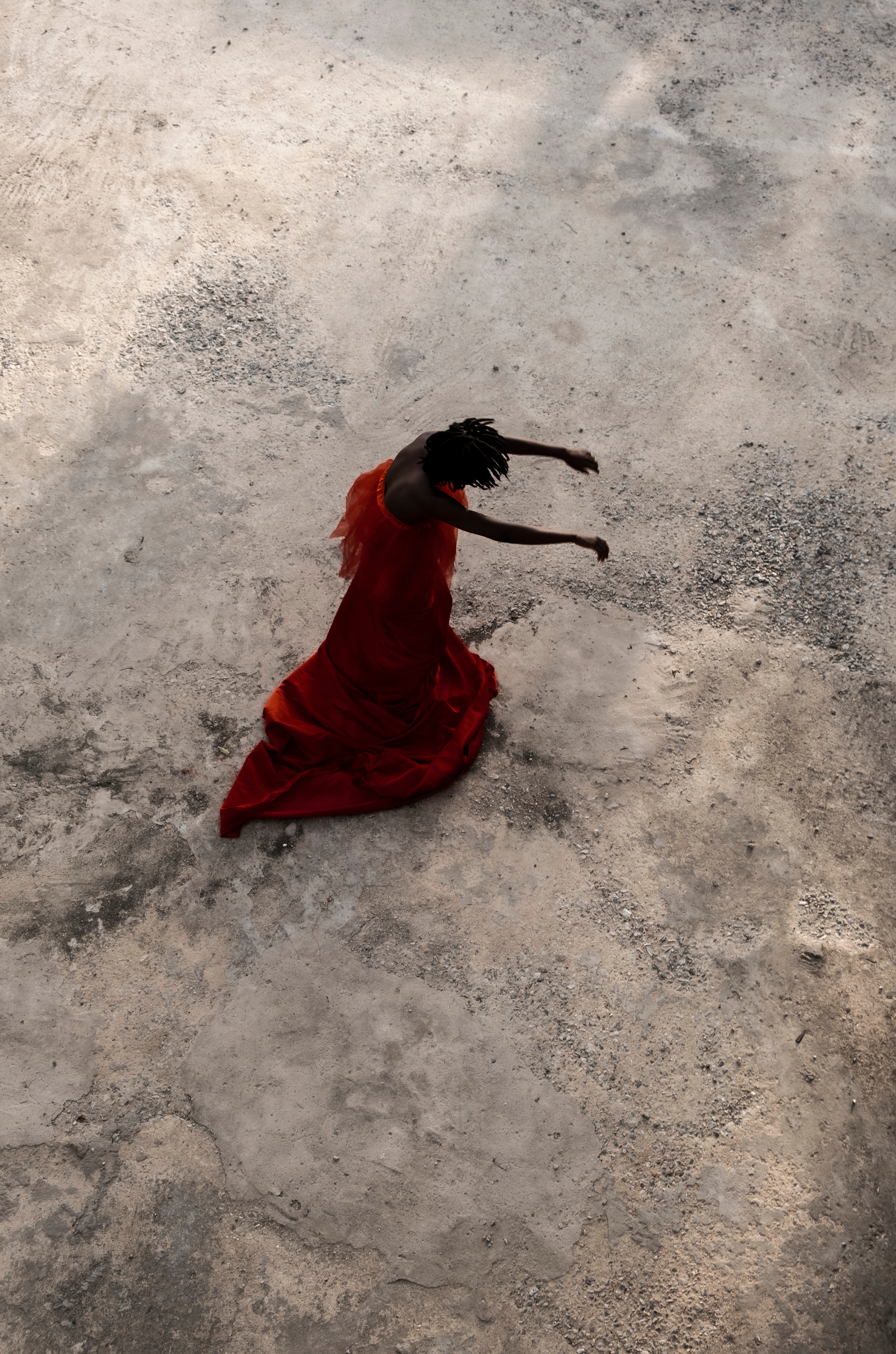 An overhead view of a person in a flowing orange dress dancing on a textured, light-colored ground.