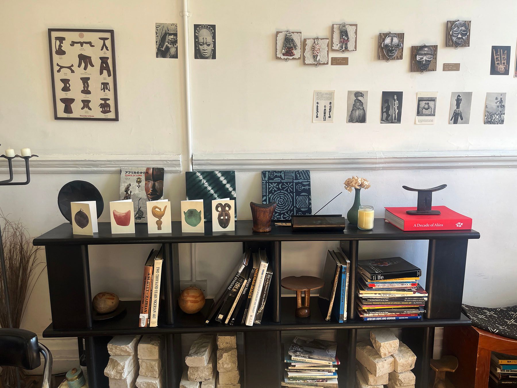 A black bookshelf displays books, greeting cards, and African-inspired decor, with a white wall above featuring various framed art and prints.