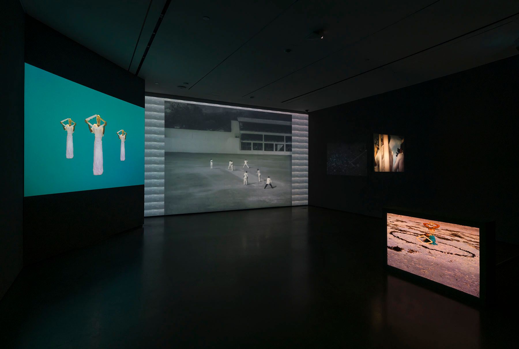 A dimly lit art gallery with multiple screens displaying video art and images, including white figures on teal, people playing cricket, abstract art, and a child drawing in sand.