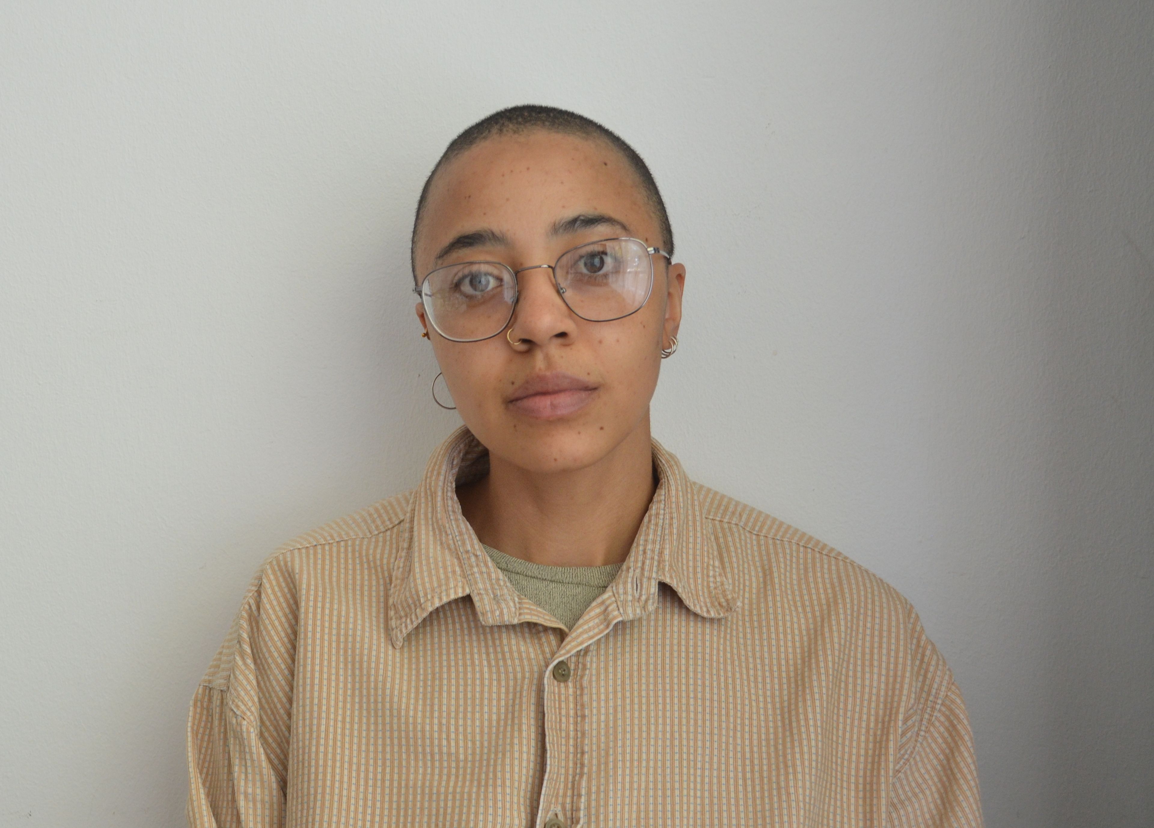 A person with a shaved head and glasses looks forward, wearing a striped tan shirt.
