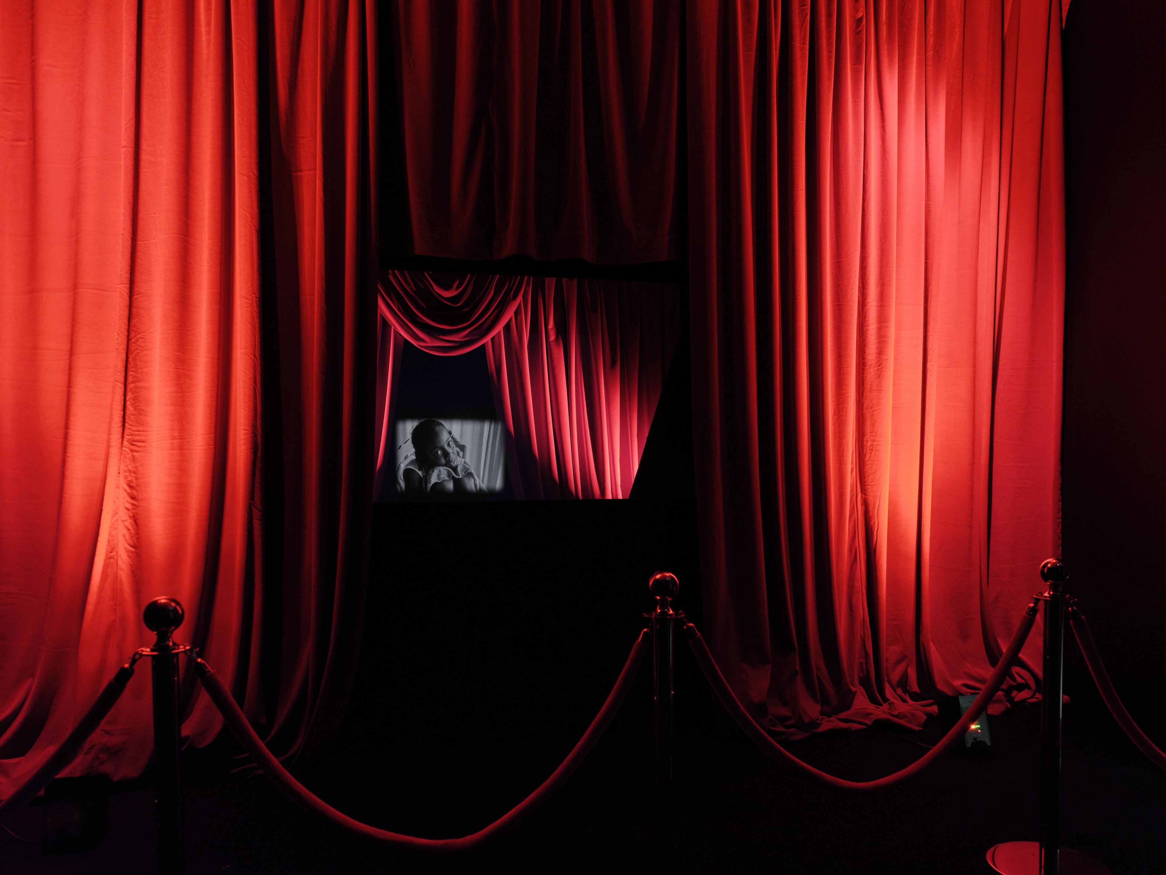 Red theatrical curtains frame a screen displaying a black-and-white image of a baby, with red velvet ropes in the foreground.