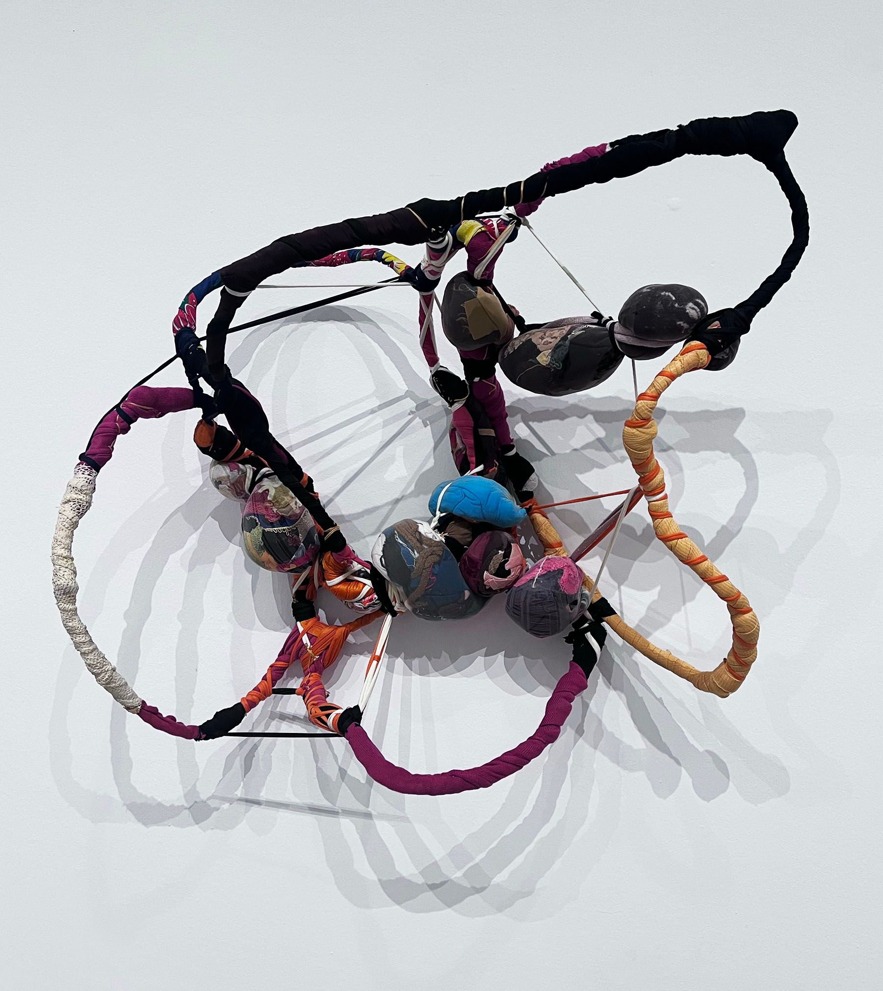 Abstract sculpture composed of multi-colored fabric-wrapped ropes and bundled forms, suspended against a white wall.