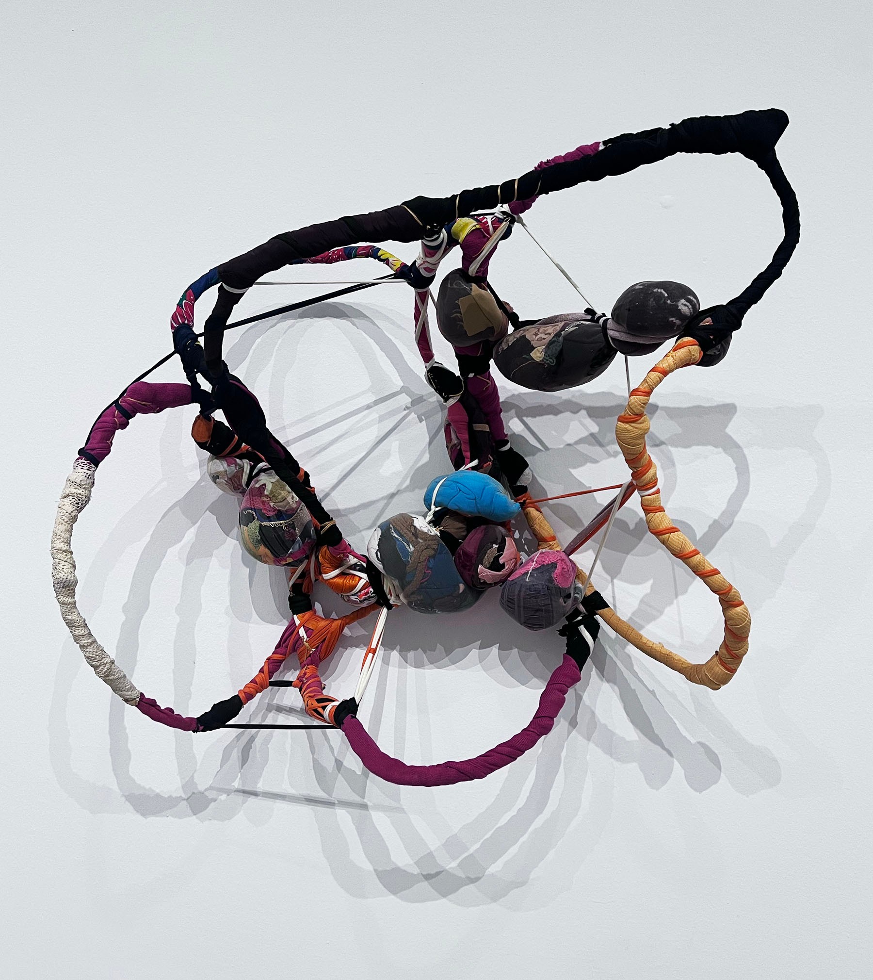 Abstract sculpture composed of multi-colored fabric-wrapped ropes and bundled forms, suspended against a white wall.