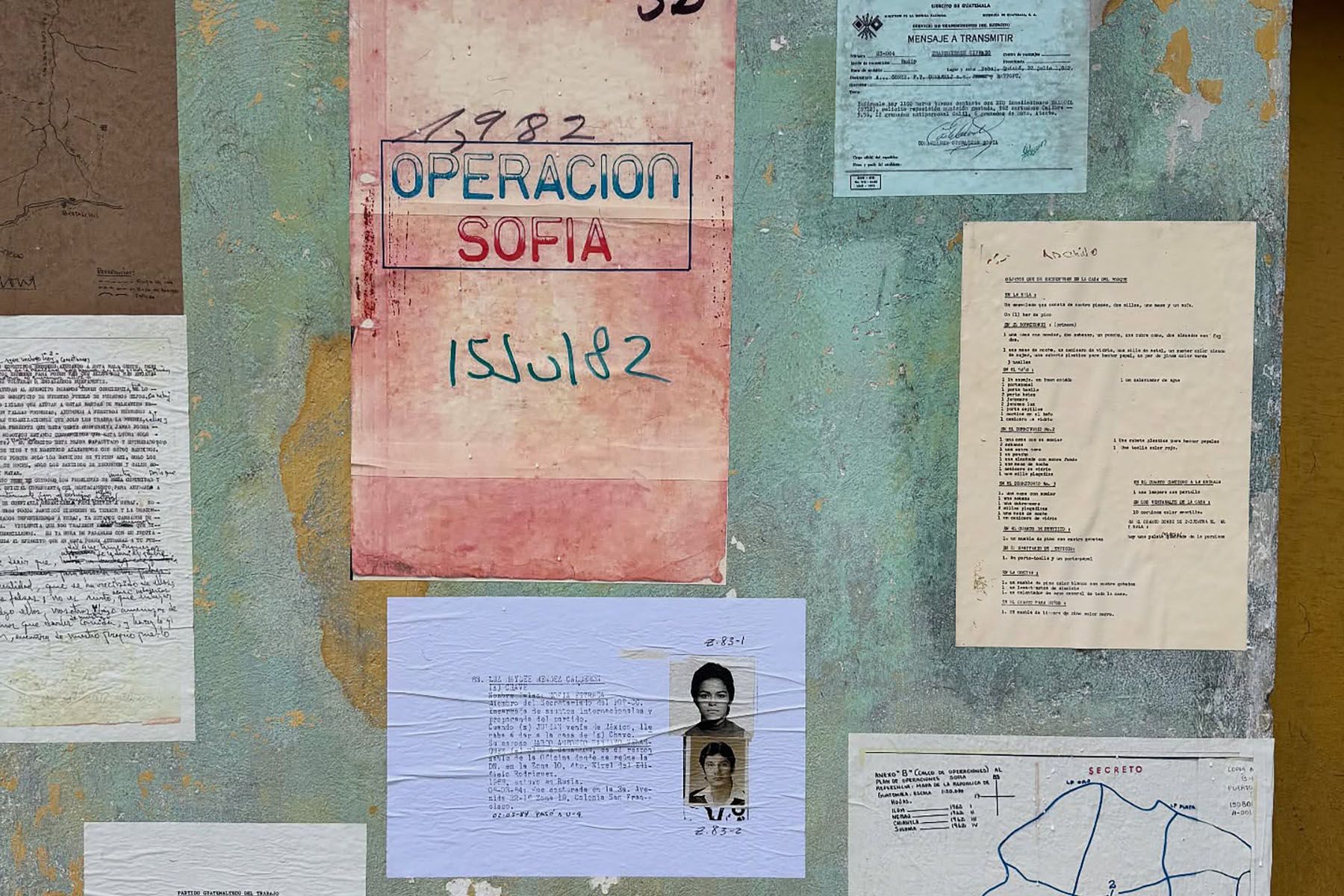 A textured wall covered with various historical documents, including one labeled "Operacion Sofia 1982" and two small passport photos.