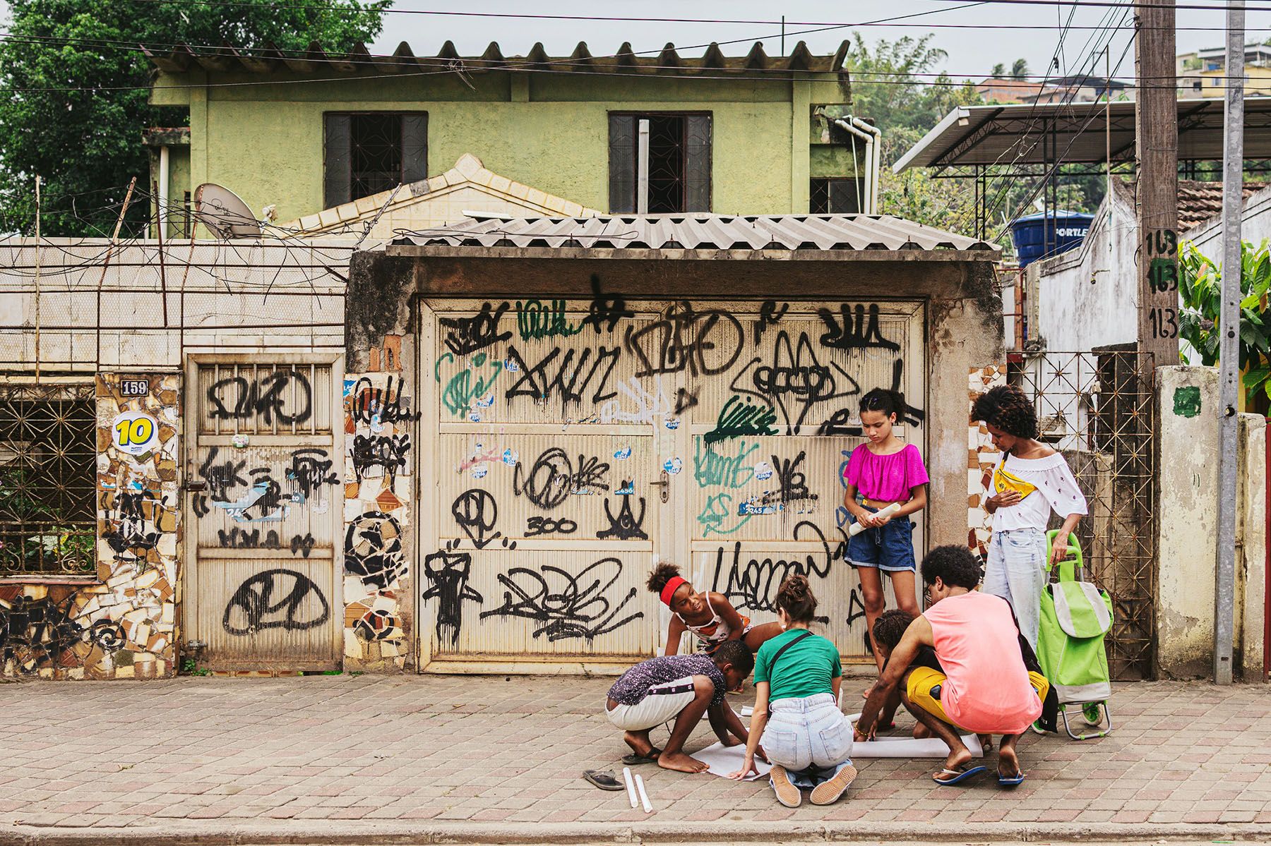 A group of young people and children gather on a sidewalk, some kneeling around a large sheet of paper, in front of a graffiti-covered gate.