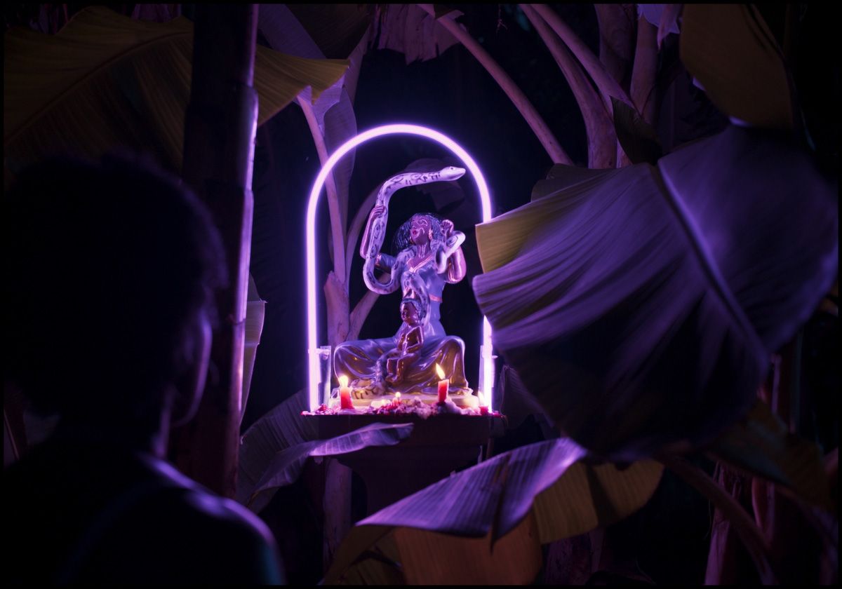 A person gazes at a purple-lit statue of a figure with a snake, framed by a neon arch and tropical leaves in the dark.