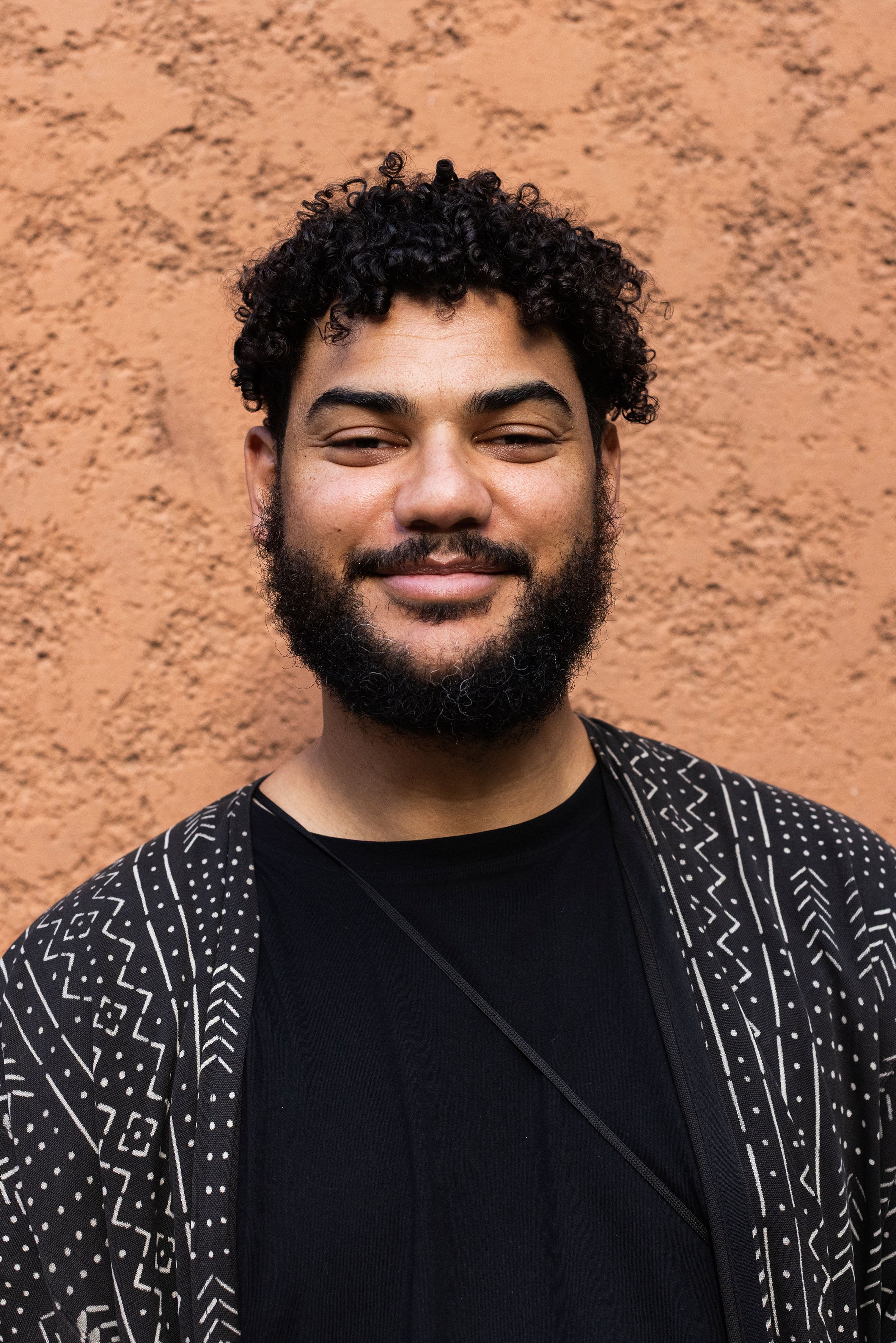 Smiling man with dark curly hair and beard in a patterned jacket against an orange wall.