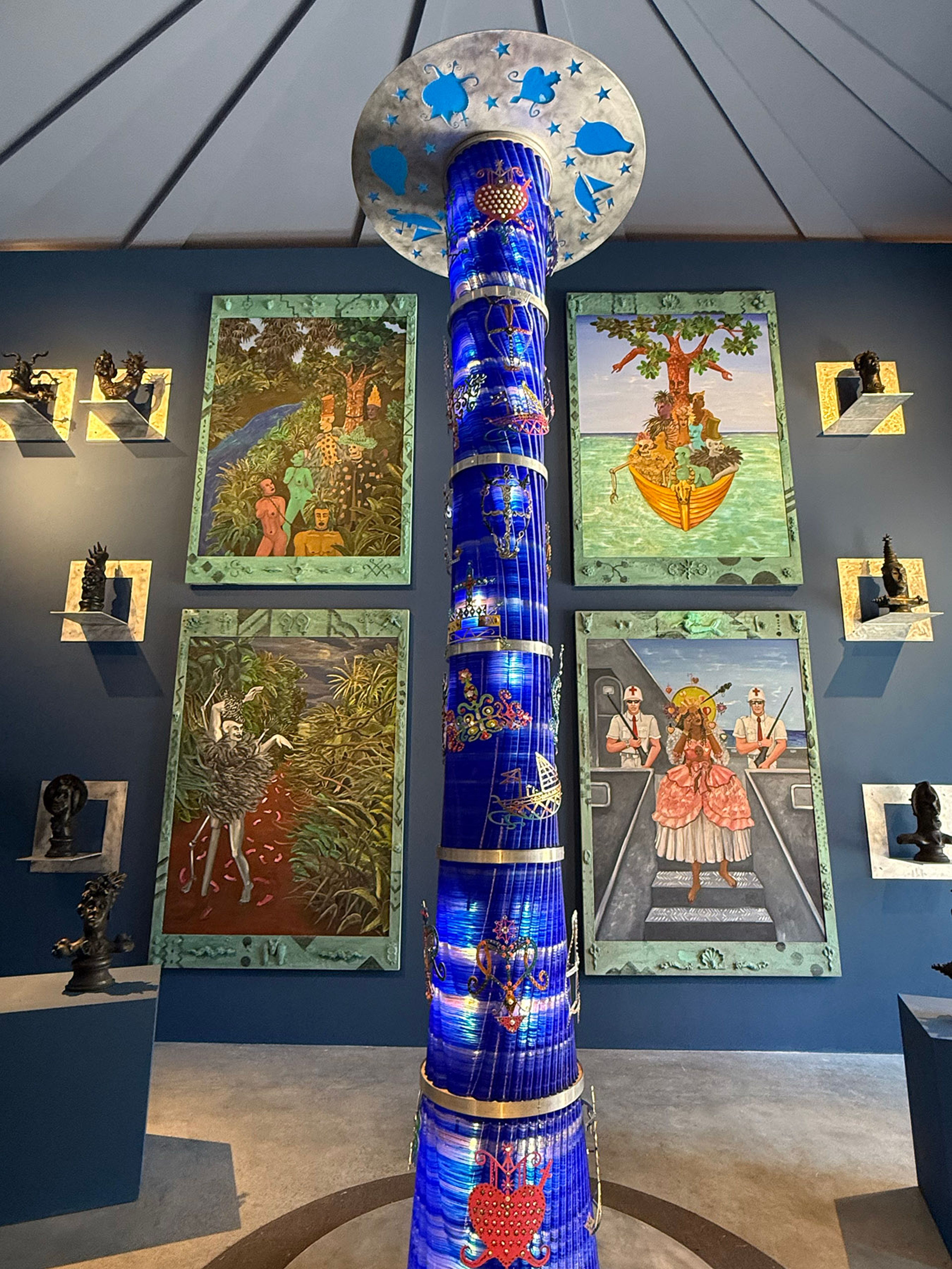 A tall, illuminated blue column with a decorated disc on top stands in a gallery, surrounded by four vibrant paintings and small dark sculptures on a blue wall.