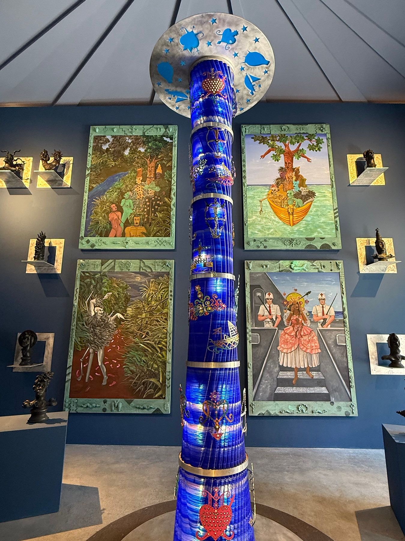 A tall, illuminated blue column with a decorated disc on top stands in a gallery, surrounded by four vibrant paintings and small dark sculptures on a blue wall.