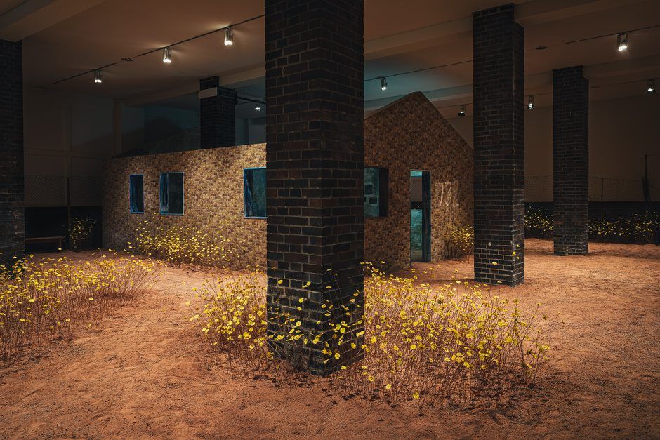 An indoor art installation with a brick house on a sandy floor dotted with yellow flowers, surrounded by dark brick columns.