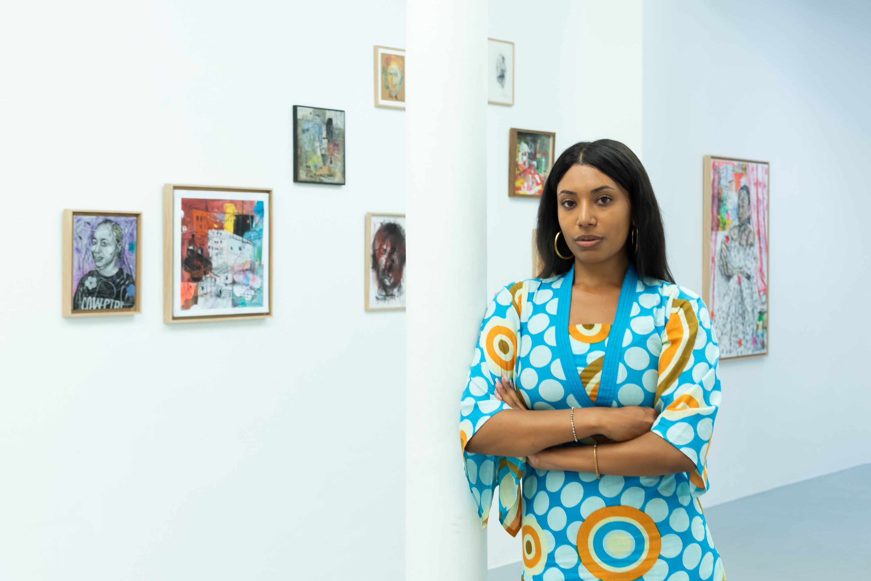 A woman in a blue, white, and orange patterned dress stands with crossed arms in an art gallery.