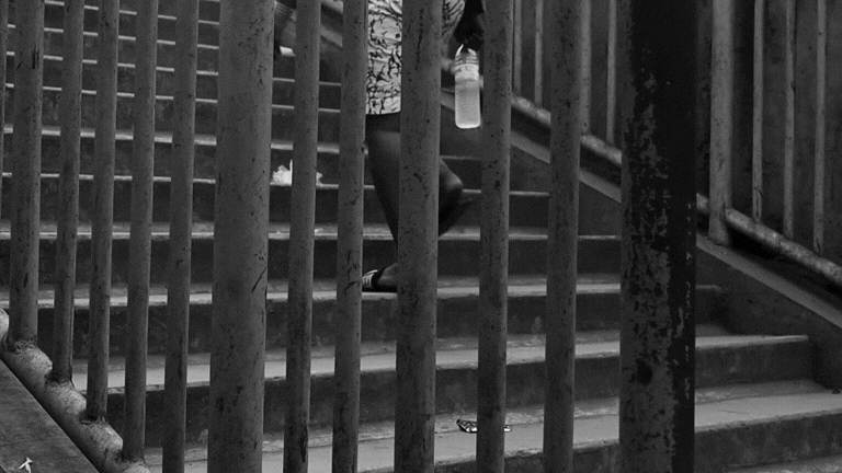 Black and white photo of a person walking up stairs behind a metal railing, holding a bottle.