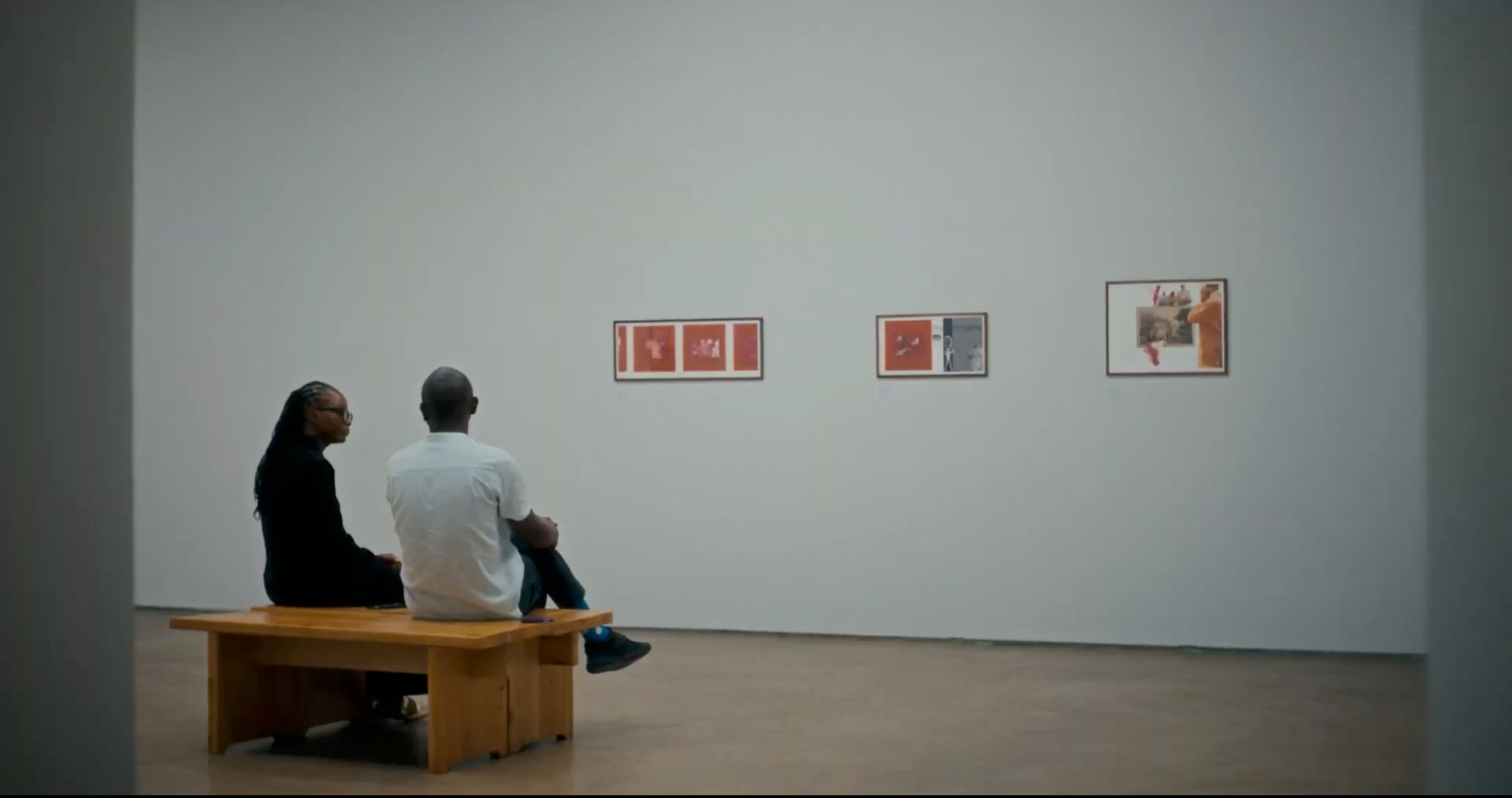 Two people sit on a bench in an art gallery, looking at three framed artworks on a white wall.