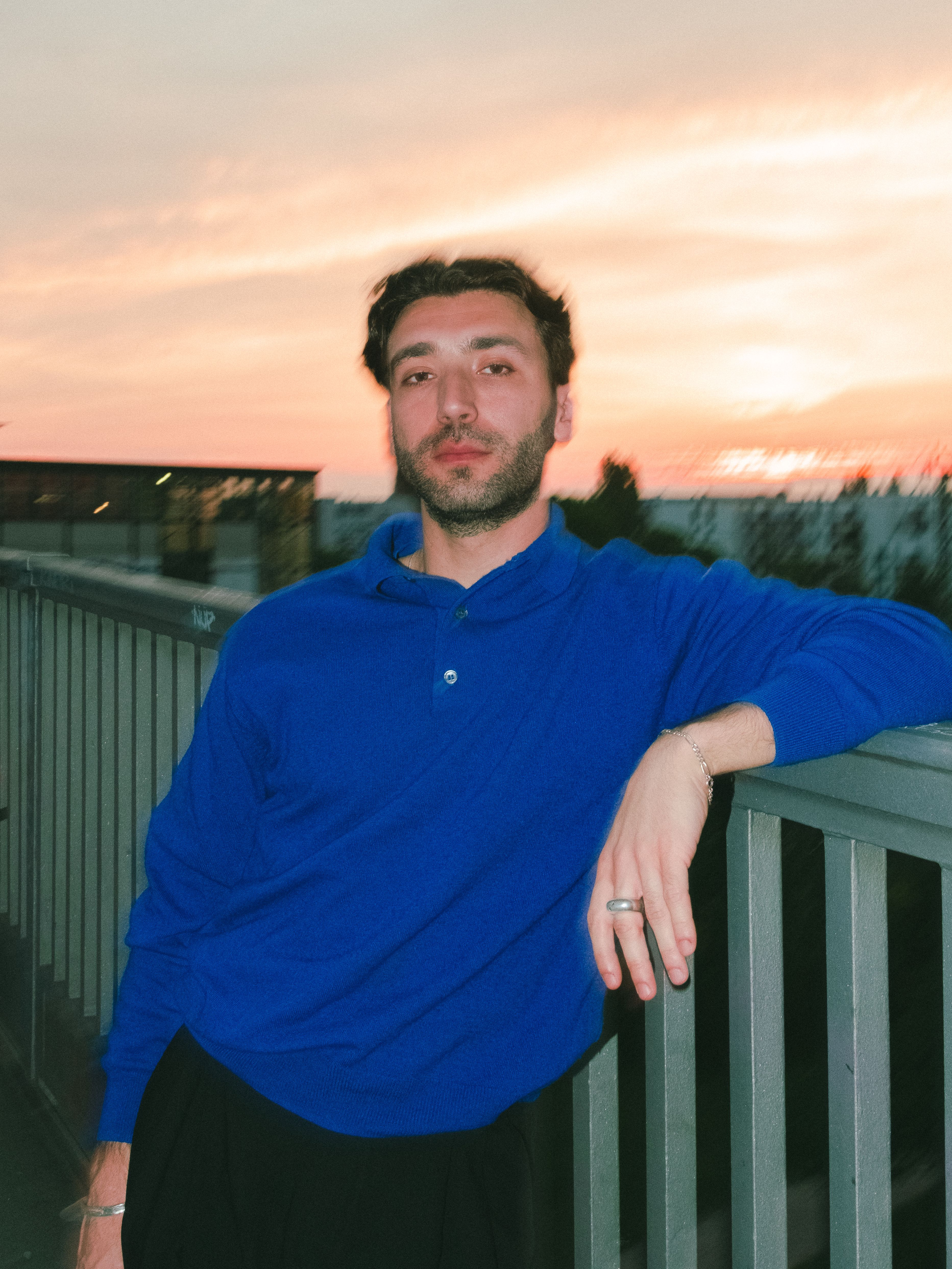 A bearded man in a blue polo shirt leans on a railing against a vibrant sunset sky.