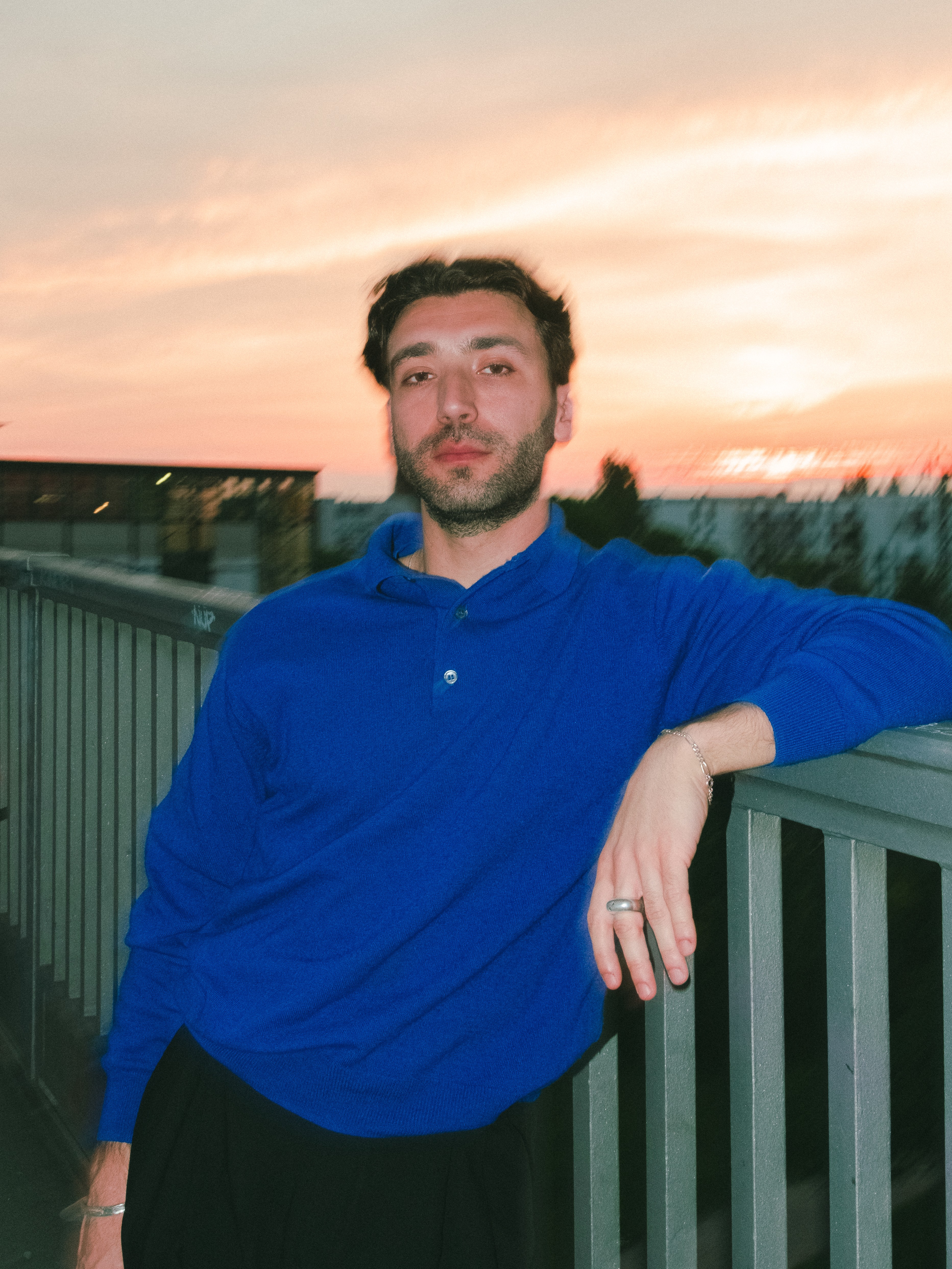 A bearded man in a blue polo shirt leans on a railing against a vibrant sunset sky.